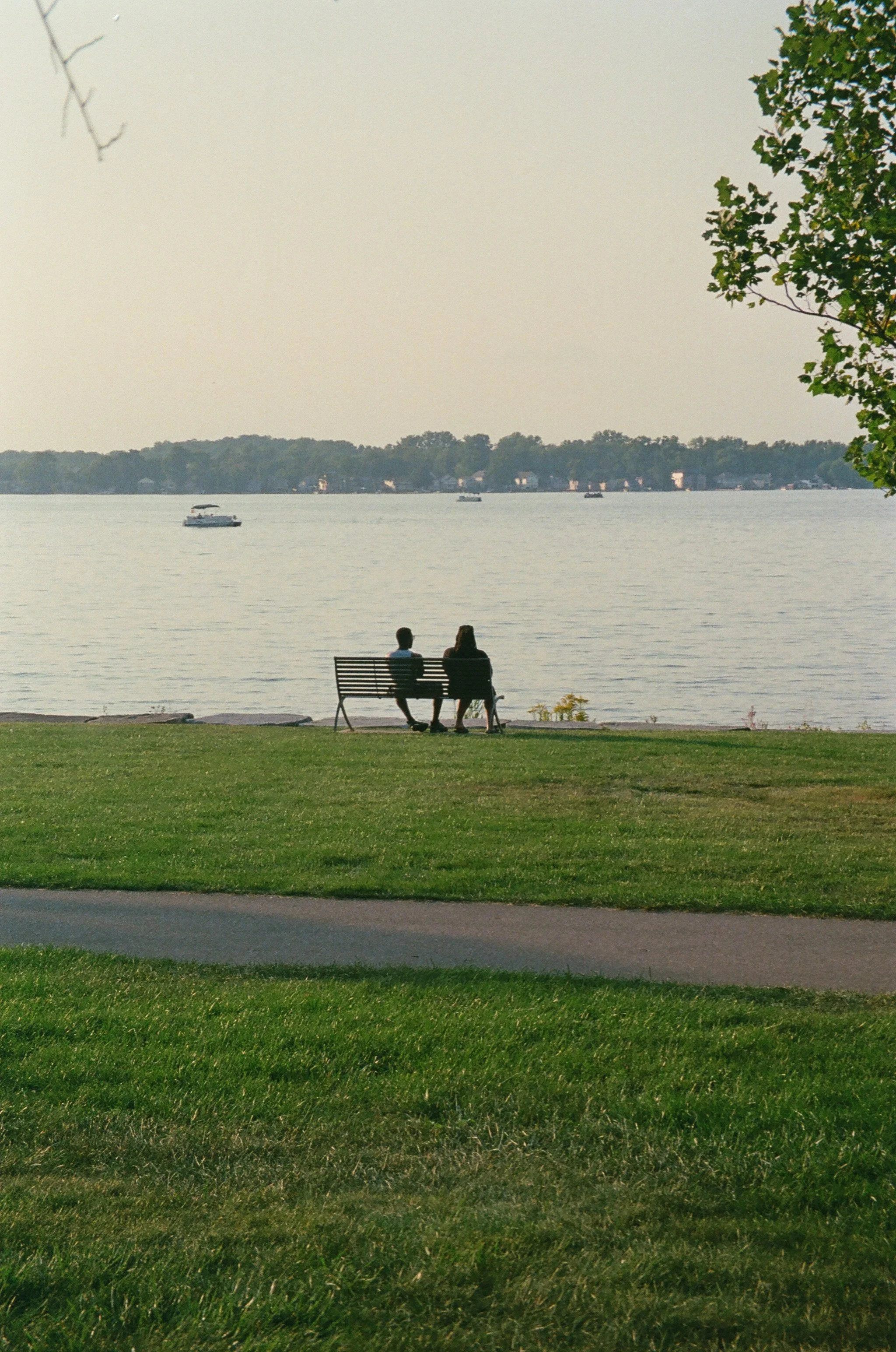 Two people sitting on a park bench by the water, looking at boats on a lake.
