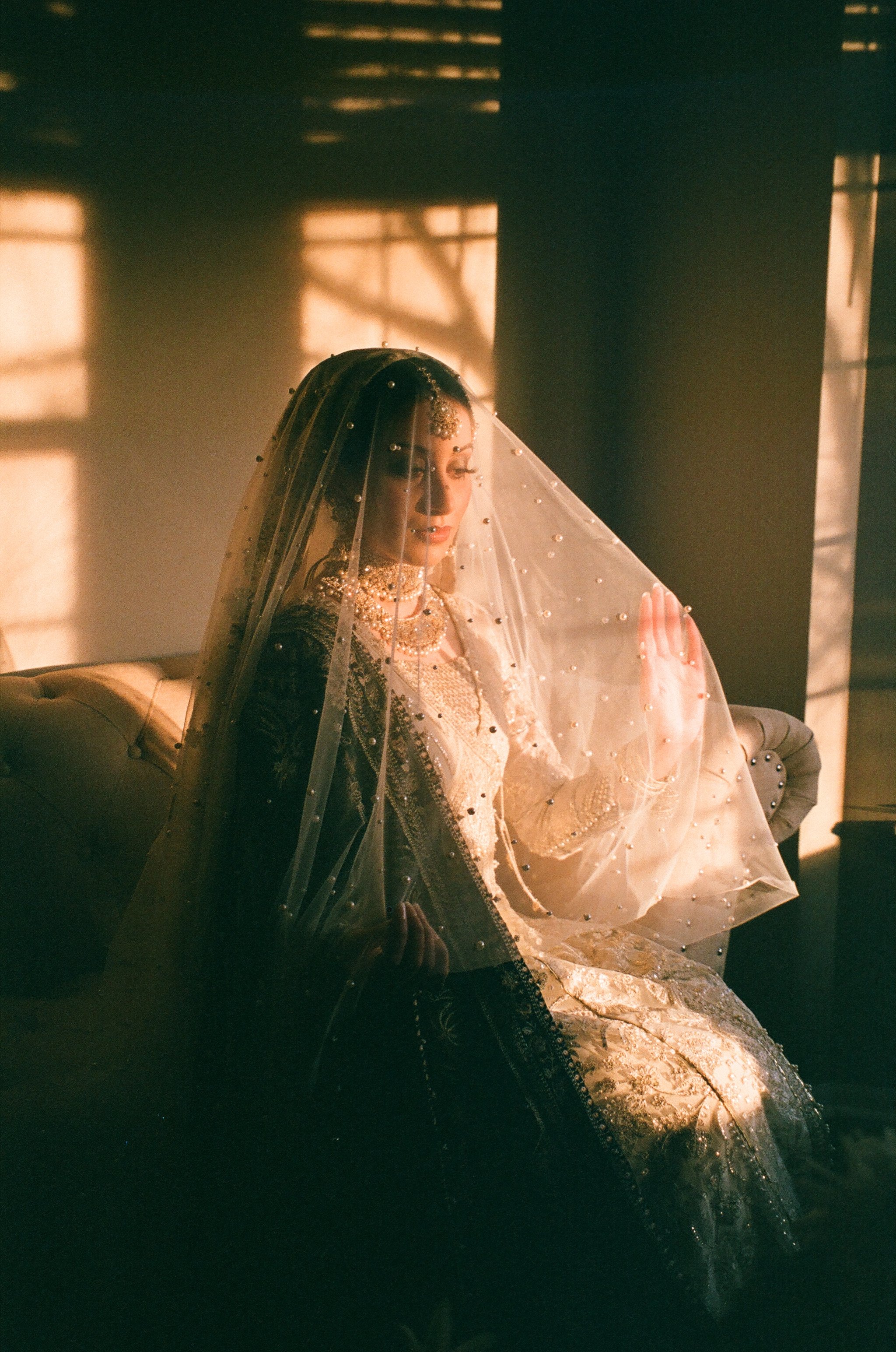 A woman dressed in traditional and ornate wedding attire, sitting on a vintage sofa, with sunlight casting shadows through window blinds.