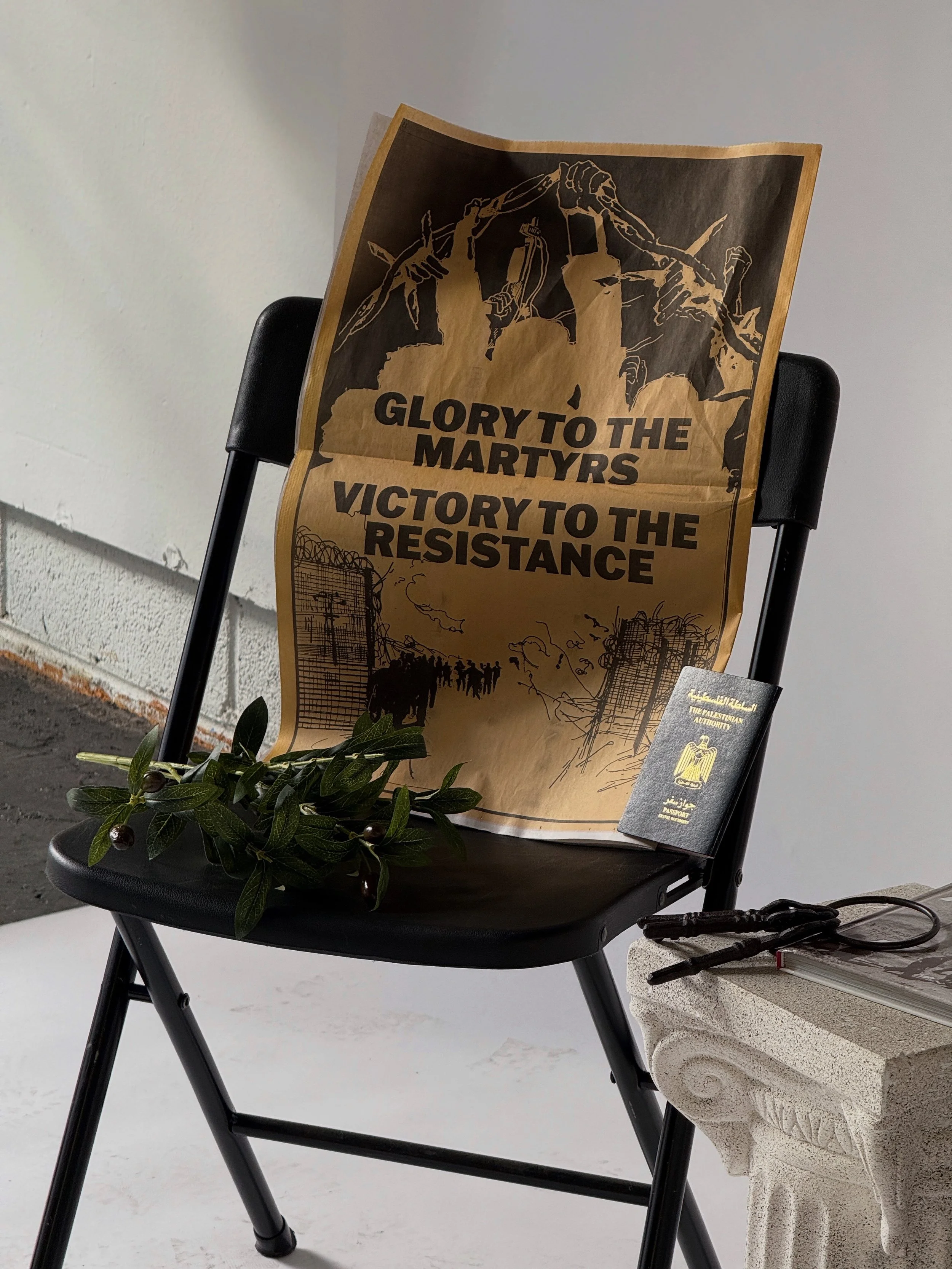 A black chair with a large poster that reads 'Glory to the Martyrs, Victory to the Resistance', a small black passport, a leafy branch, and some black strings on the armrest. The chair is next to a stone pillar and against a plain gray wall.