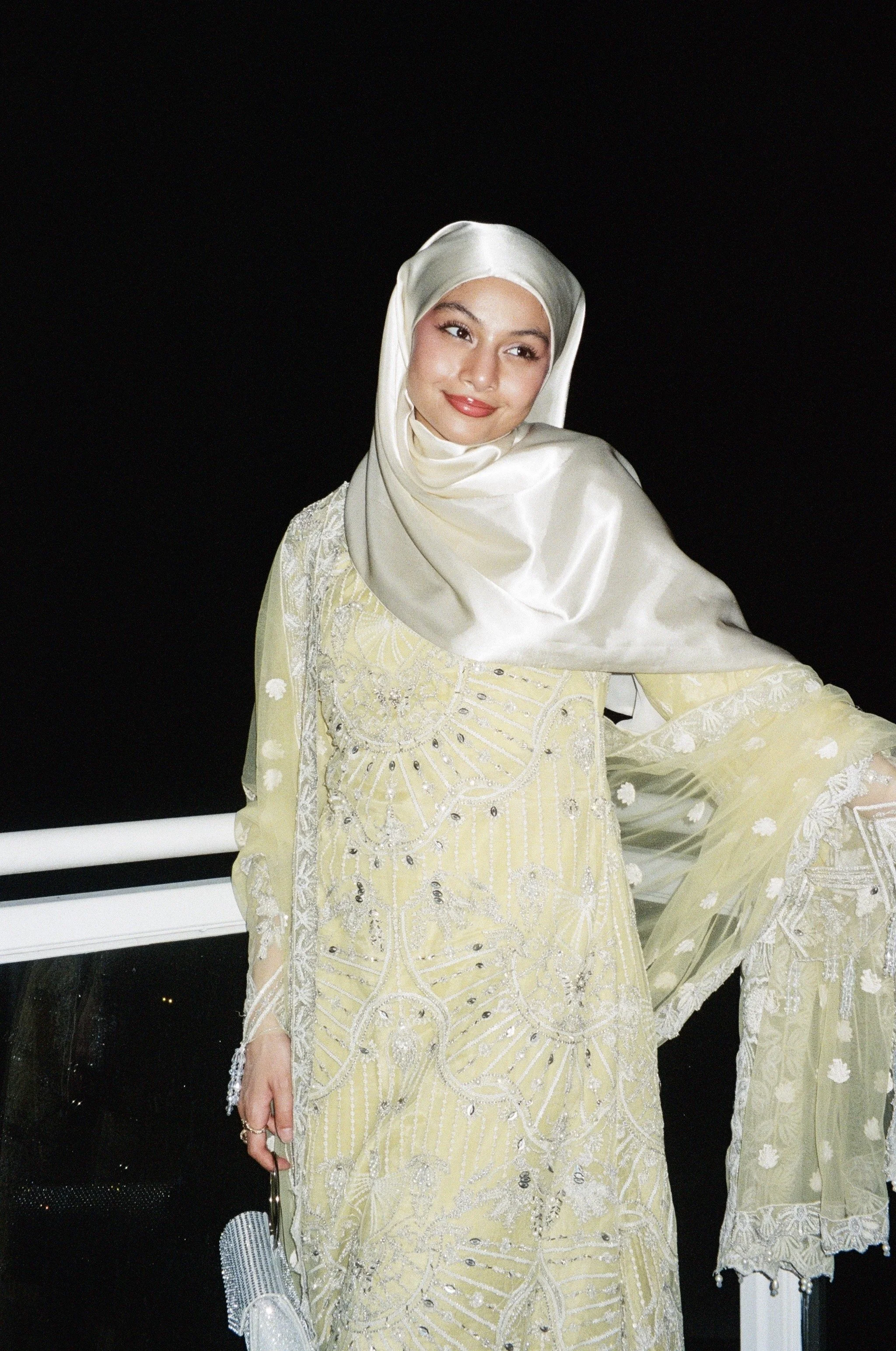 A woman in an elegant yellow dress with intricate embroidery, wearing a cream-colored satin hijab, standing outdoors at night.