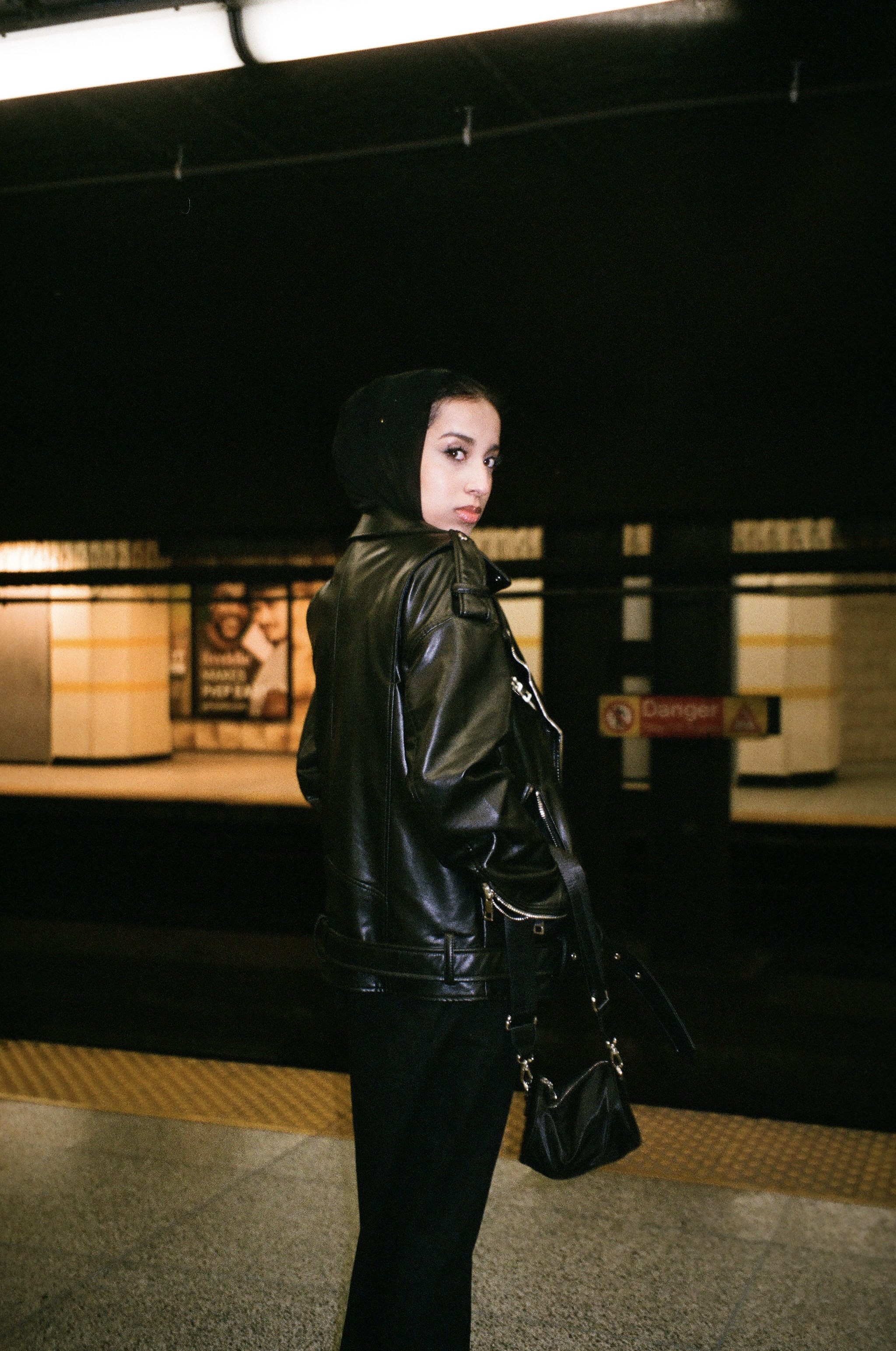 A woman in a black leather jacket and black pants stands on a train station platform at night, looking over her shoulder.