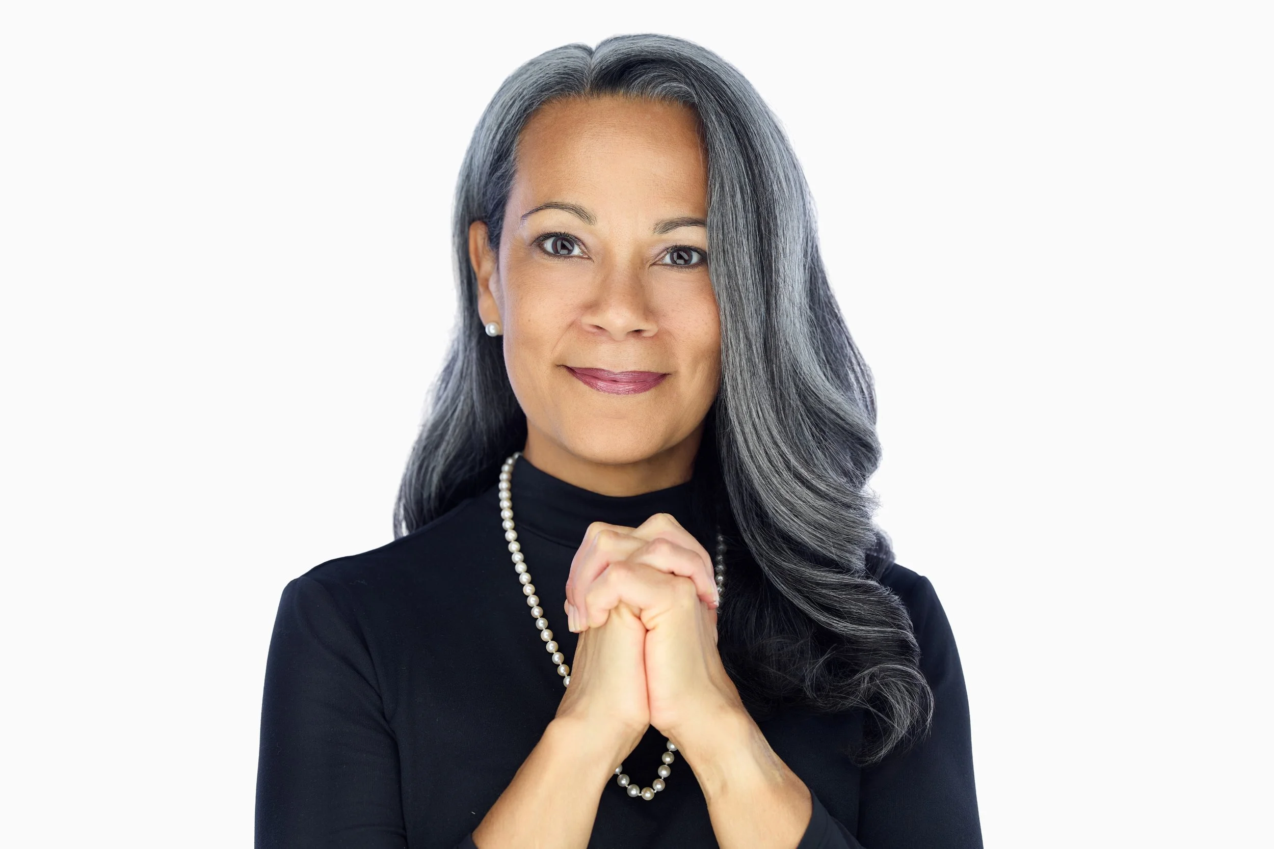 Portrait of a mature woman with long gray hair, wearing a black top and pearl necklace, holding her hands together in front of her chest, smiling softly against a plain white background.