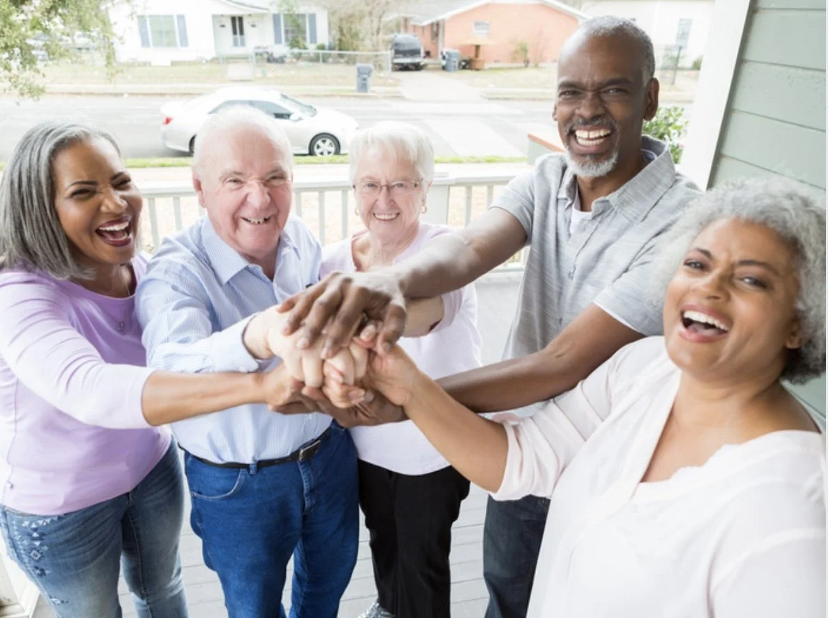 Six diverse people stand together on a porch, smiling and laughing while placing their hands together in a team gesture.