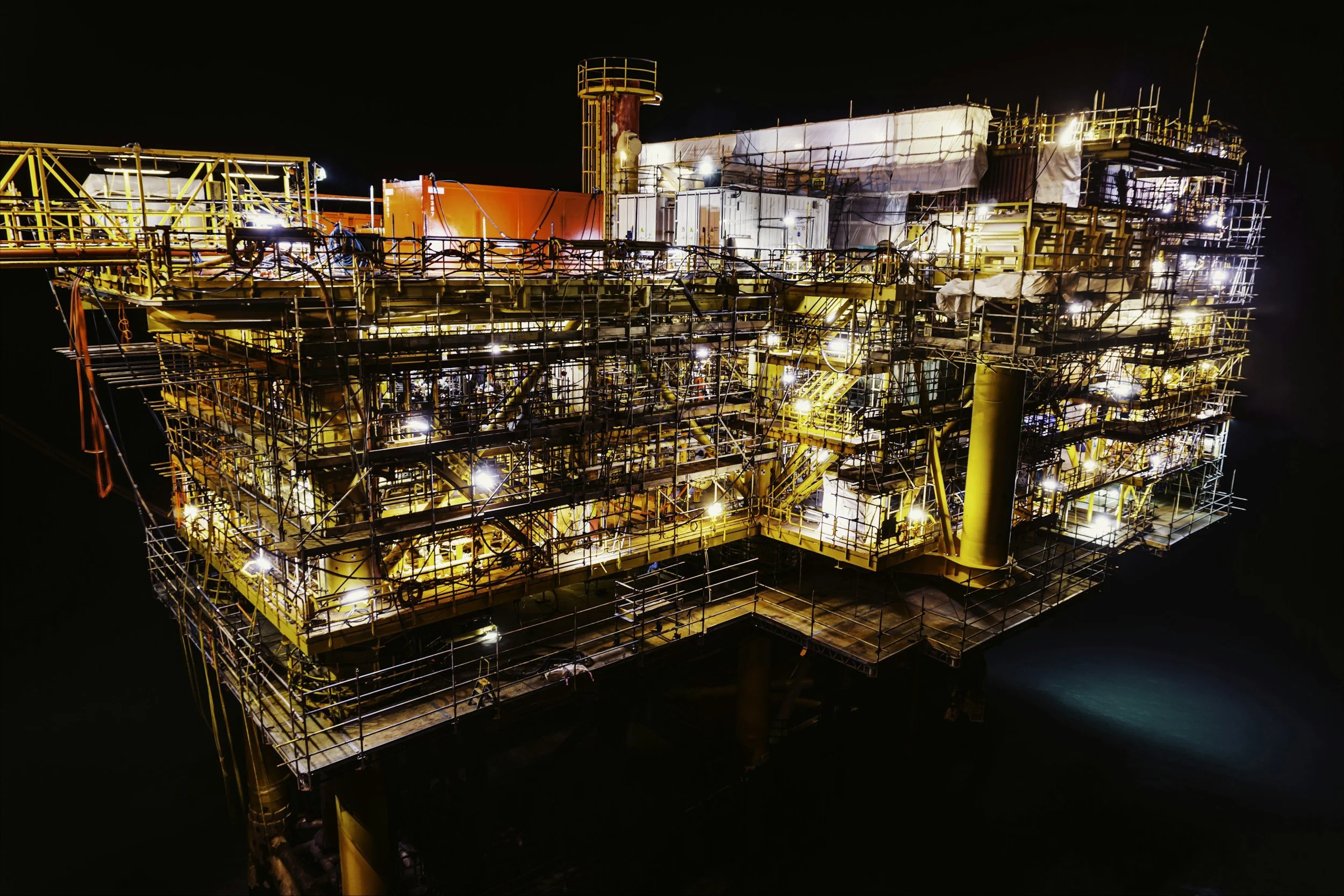Nighttime view of an offshore oil platform illuminated with bright lights, showing scaffolding and structural components.