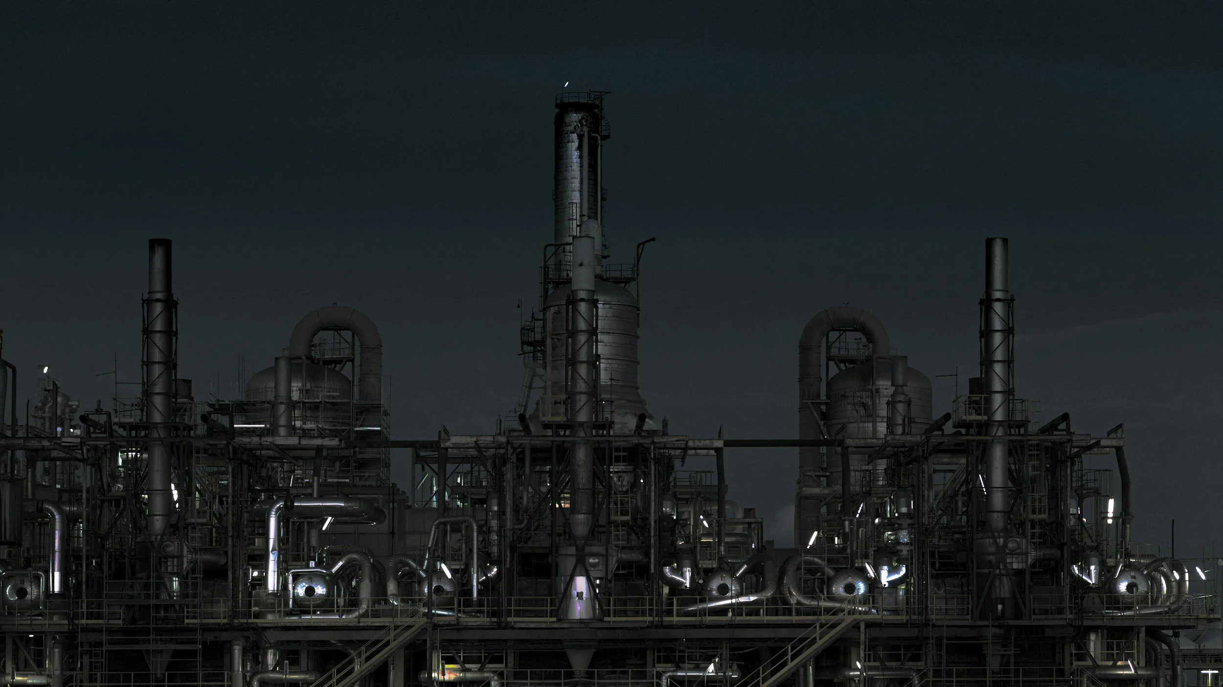 Nighttime view of an industrial refinery with multiple steel pipes and tall towers illuminated by small lights.