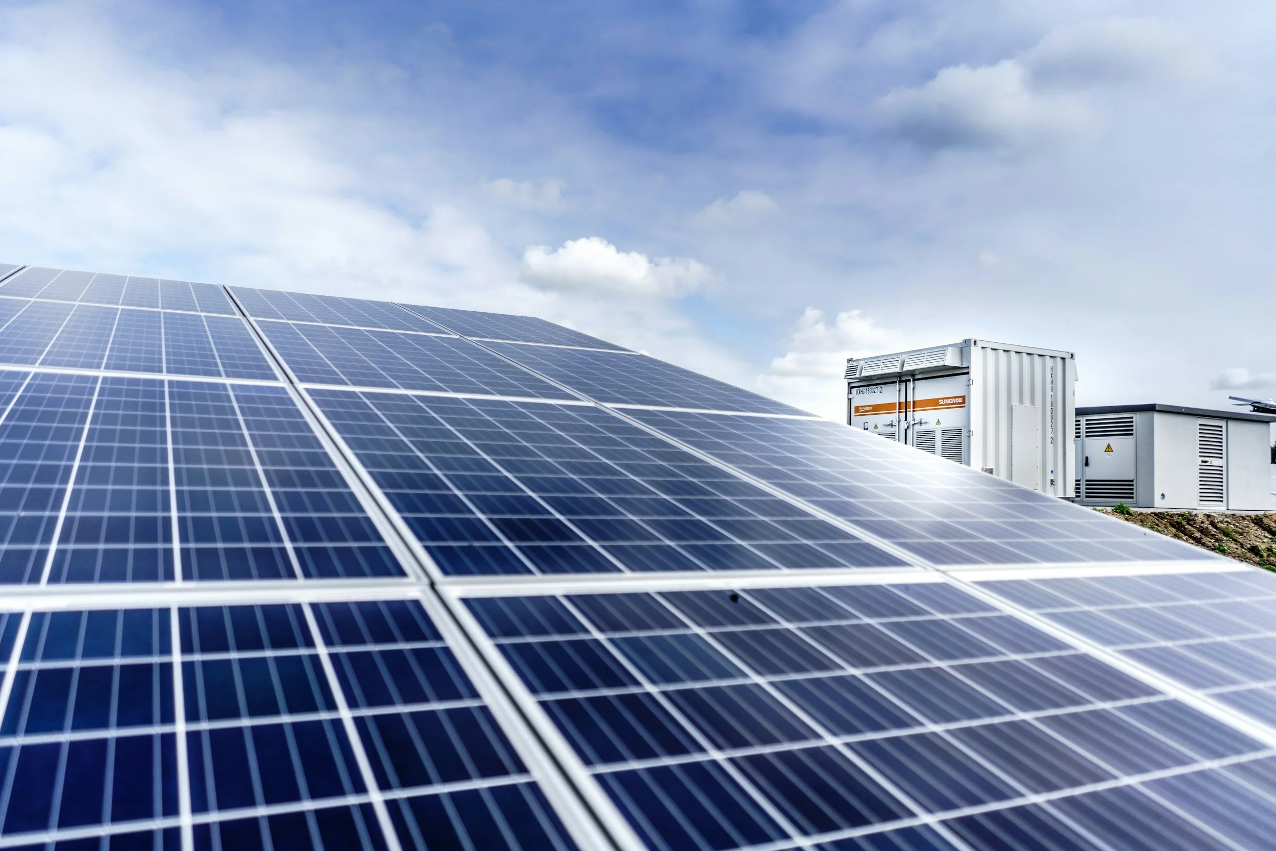 Close-up of solar panel array with utility equipment in the background under a partly cloudy sky.