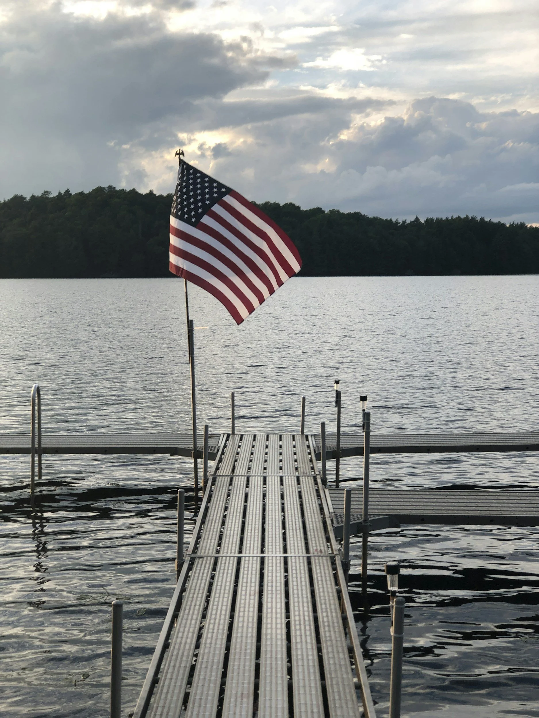 American flag on a dock overlooking a lake with a tree-lined shoreline in the distance and a cloudy sky overhead.