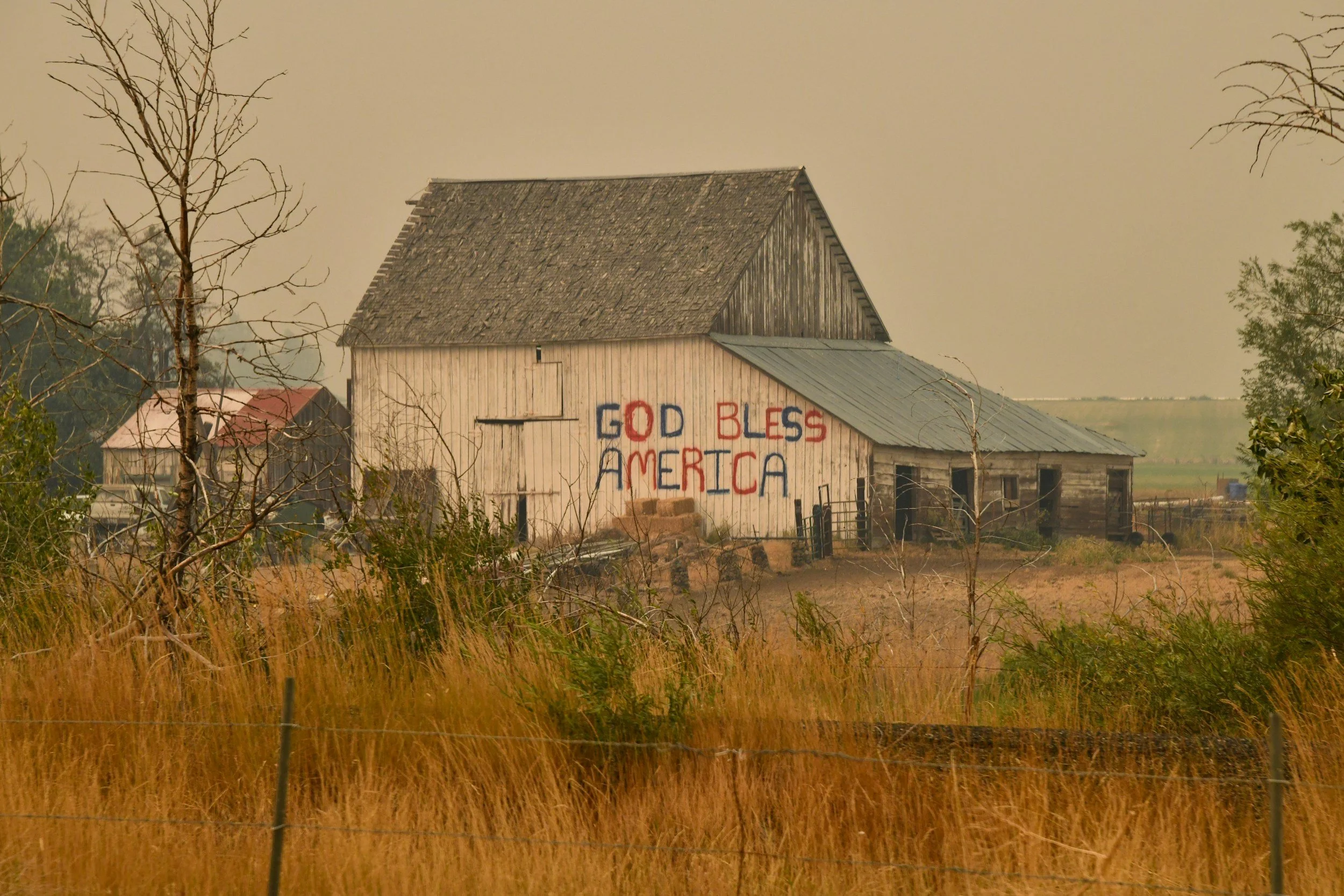 Old barn on a farm with the words 'God Bless America' painted on the side in red, white, and blue paint, surrounded by dry grass and leafless trees.