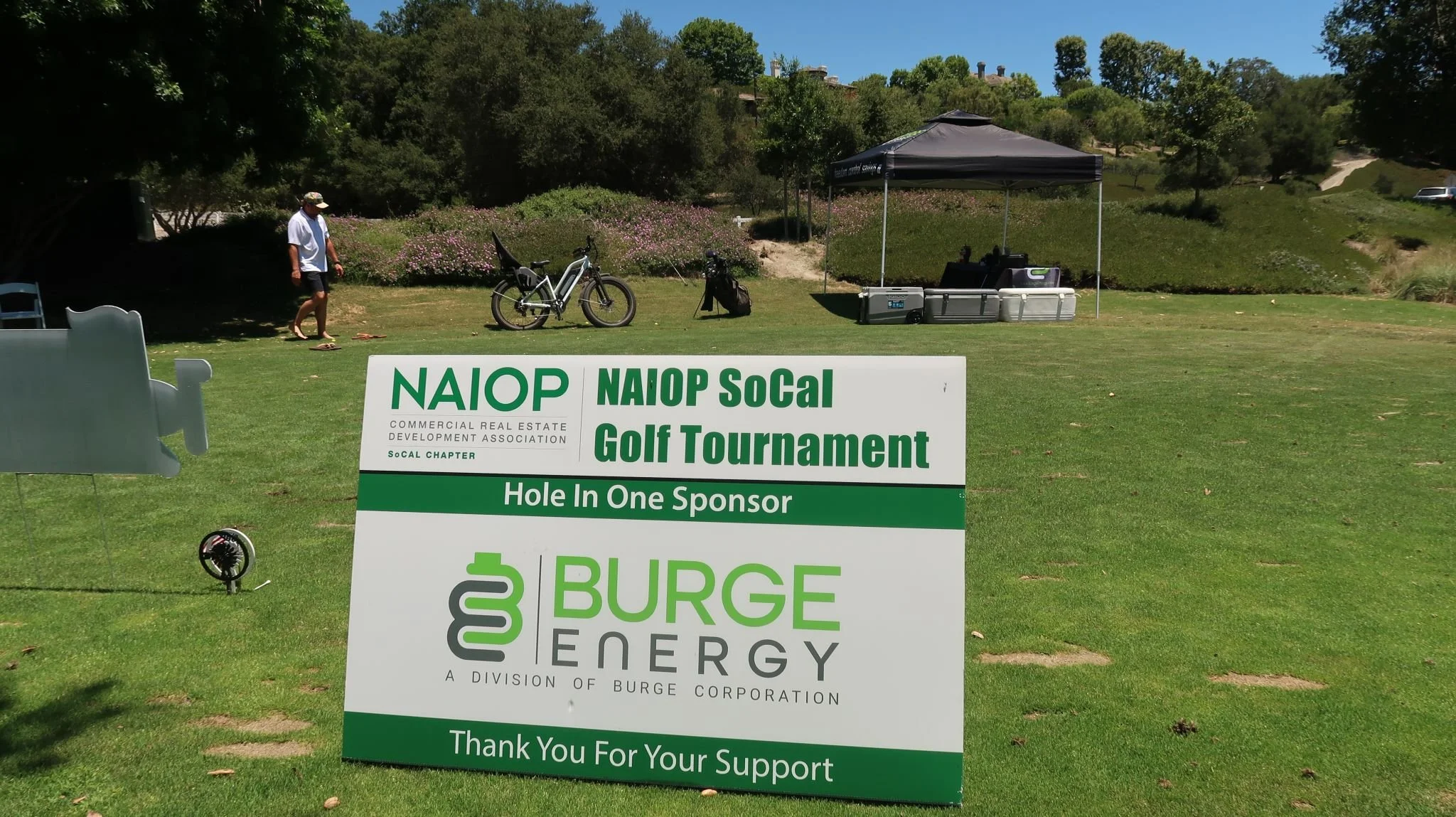 A golf course with a man walking on the grass near a bicycle and a sponsor sign for the NAIOP SoCal Golf Tournament, sponsored by Burge Energy, under a tent with cooler boxes, with trees and a hill in the background.