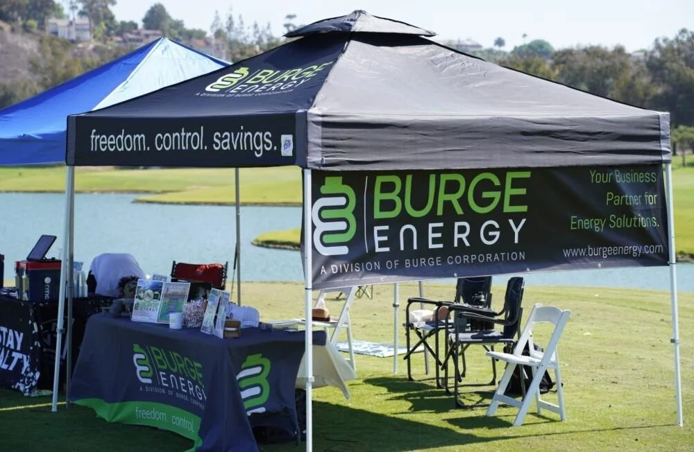 A large outdoor booth with a black and green Burge Energy tent on a grassy area near a lake, with chairs and promotional material displayed.