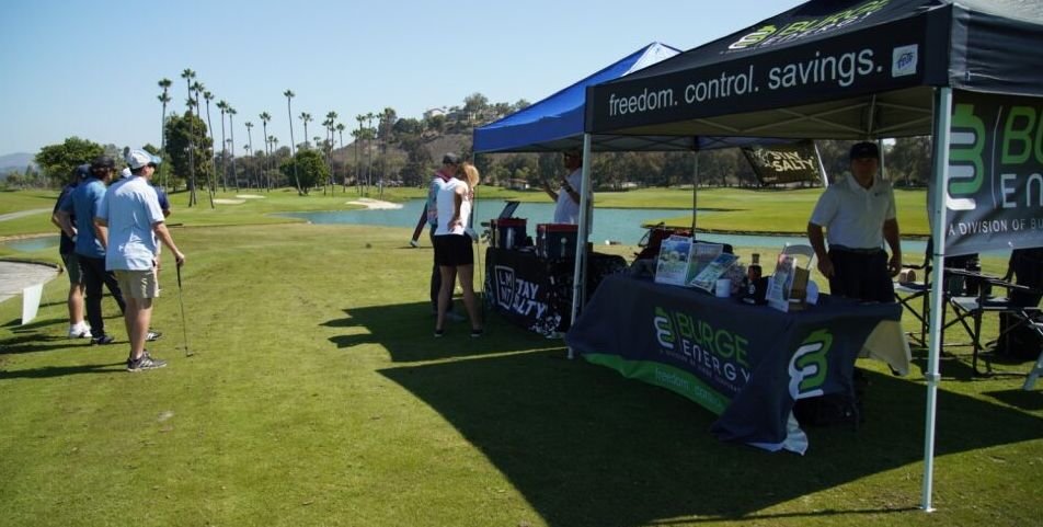 Golf course with people playing, promotional tents for Burge Energy and Toyota, and palm trees in the background.