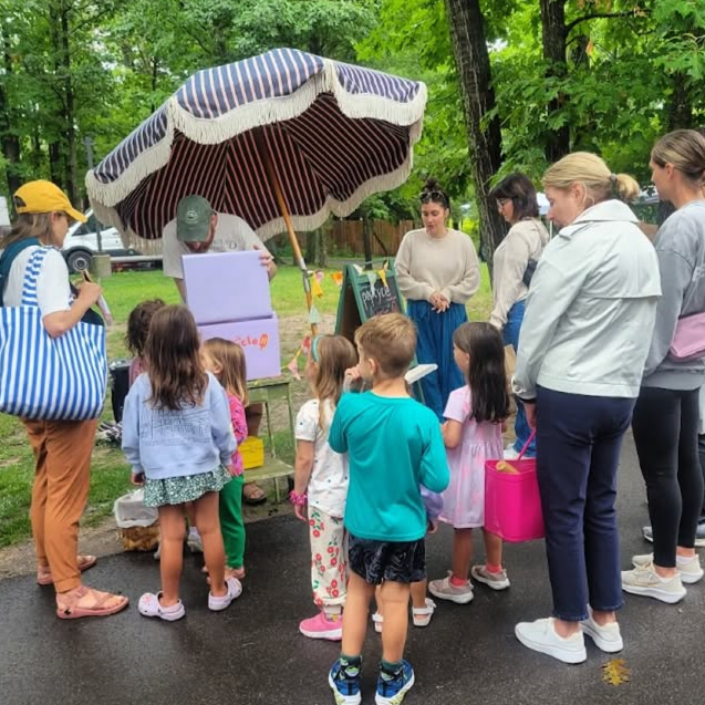 Children and adults gather outdoors around a vendor stand with an umbrella, at a park or fair, listening to a man who is preparing something.