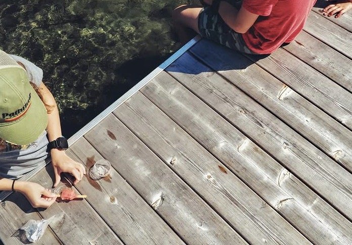 Two children sitting on a wooden dock by a body of water, one holding a fishing pole and the other looking at the water.