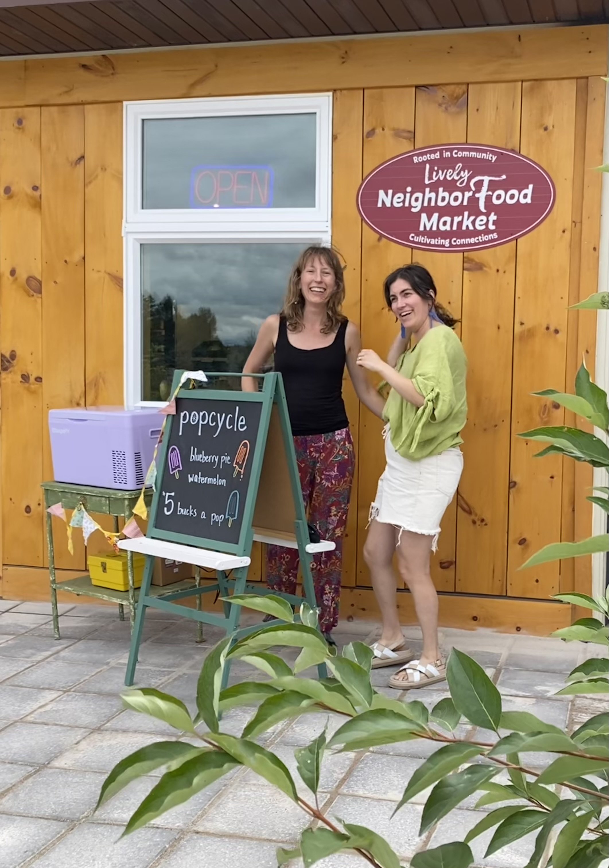 Two women standing outside a neighborhood food market with an orange wooden wall behind them. One woman is wearing a black tank top and patterned pants, the other is in a yellow-green shirt and white shorts. There is a sign in front of them advertising popsicles and the market has an