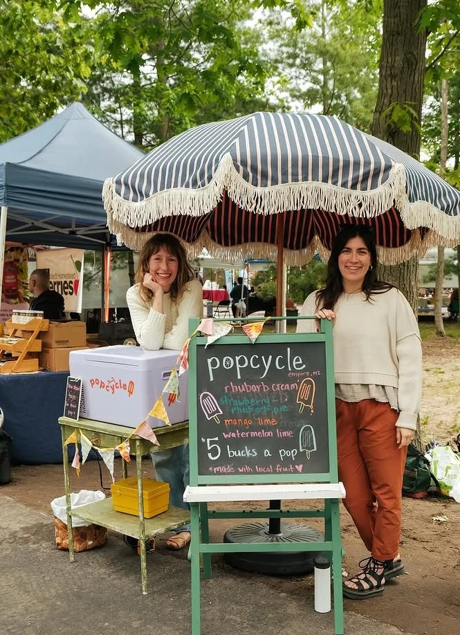 Two women at a roadside stand with a striped umbrella. The stand sells popsicles and features a chalkboard sign listing flavors and price.
