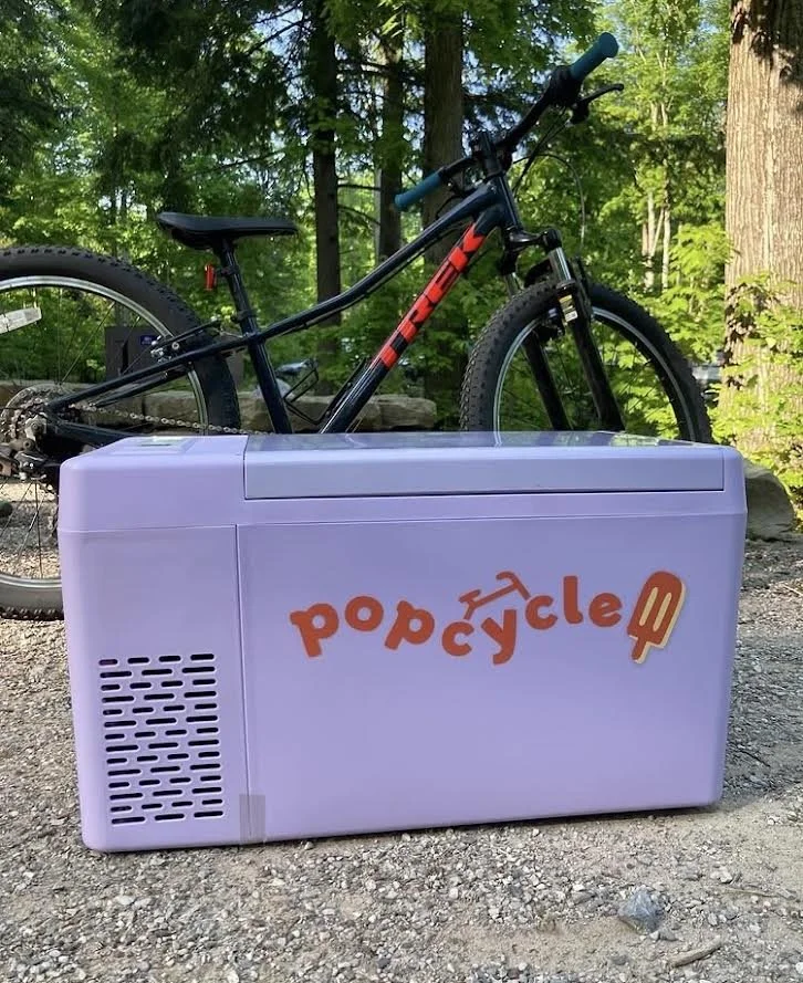 A purple freezer with the word 'popcycle' written in orange and decorated with a popicles icon. Behind the freezer, there is a mountain bike with a black frame and red accents, set outdoors in a wooded area.
