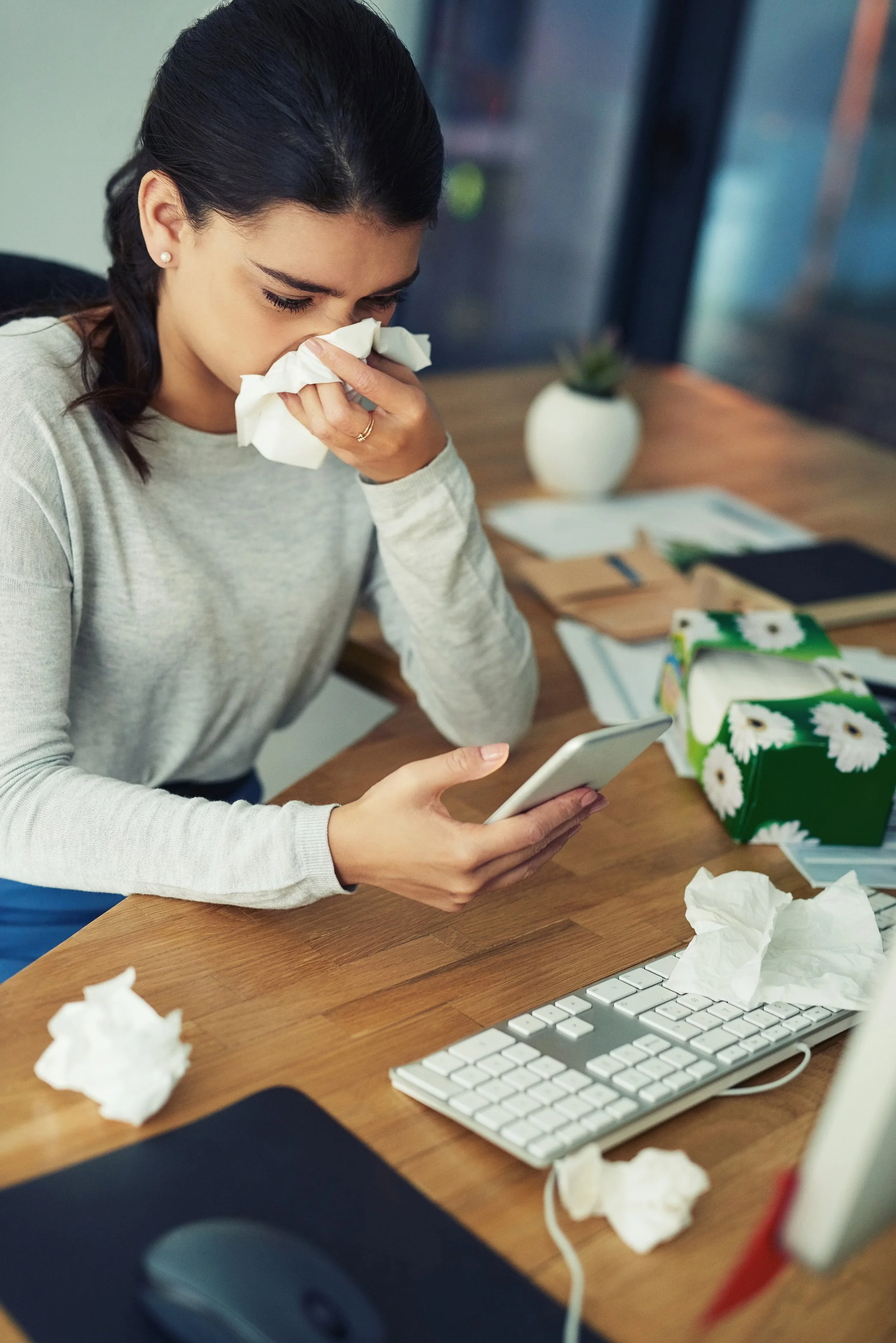 Woman sneezing into tissue while looking at her phone at a cluttered desk with crumpled tissues, a keyboard, and other office supplies.