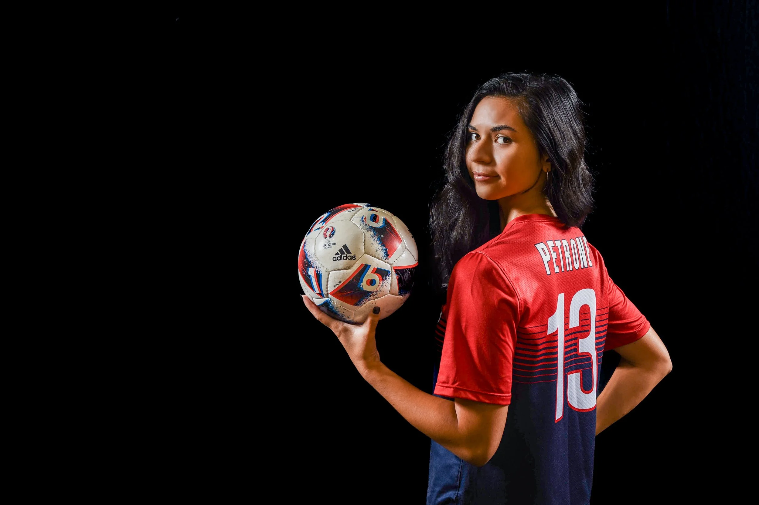 A woman in a red and navy soccer jersey holding a soccer ball, standing against a black background.