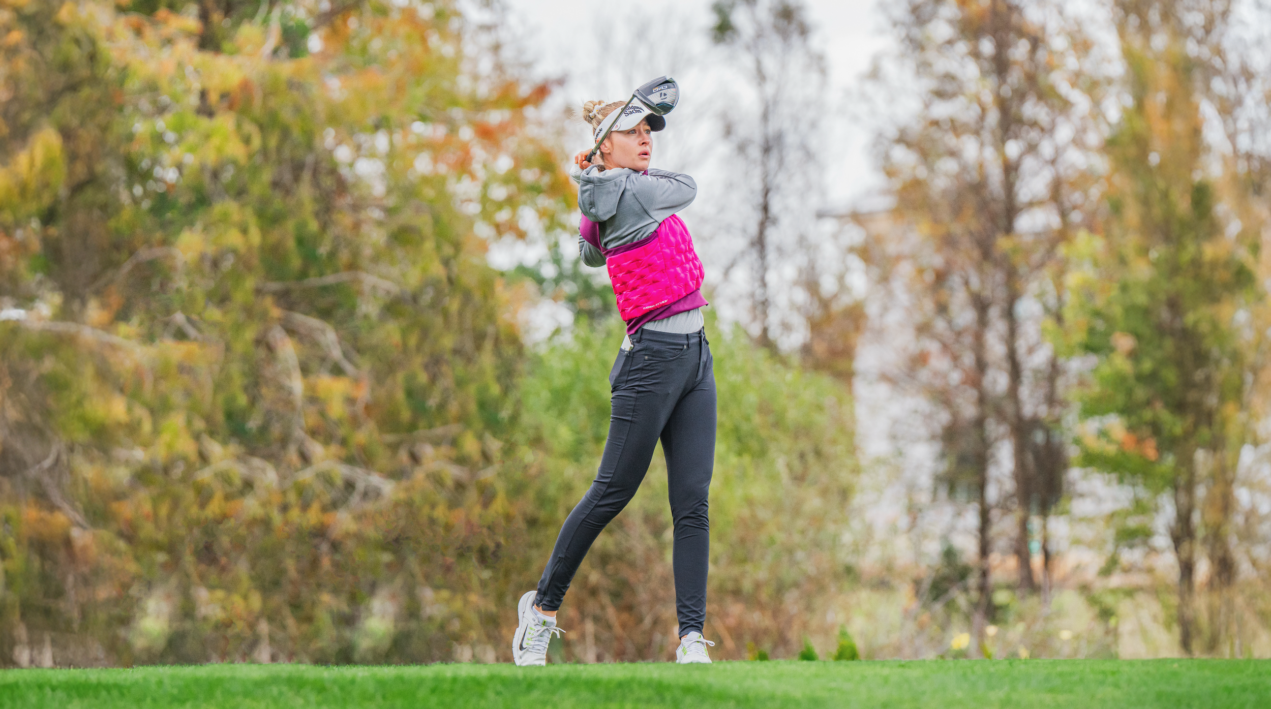 Nelly Korda playing golf on a grassy course with trees in autumn foliage in the background, holding a golf club over her shoulder. PNC Championship. 
