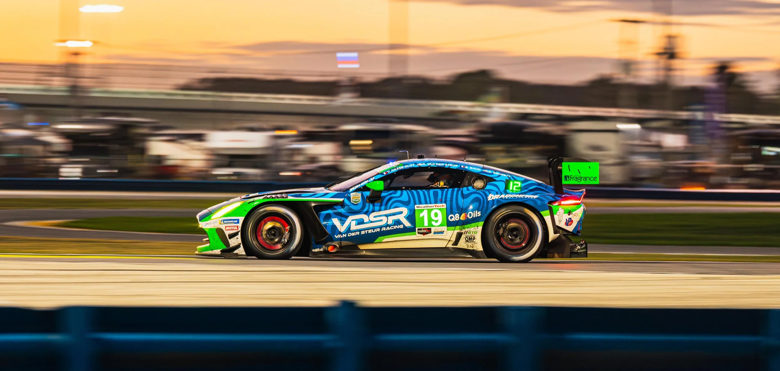 A race car speeds along a track at sunset, with a background of blurred spectators and structures. The car is blue with green accents, displaying sponsor logos and the number 19.