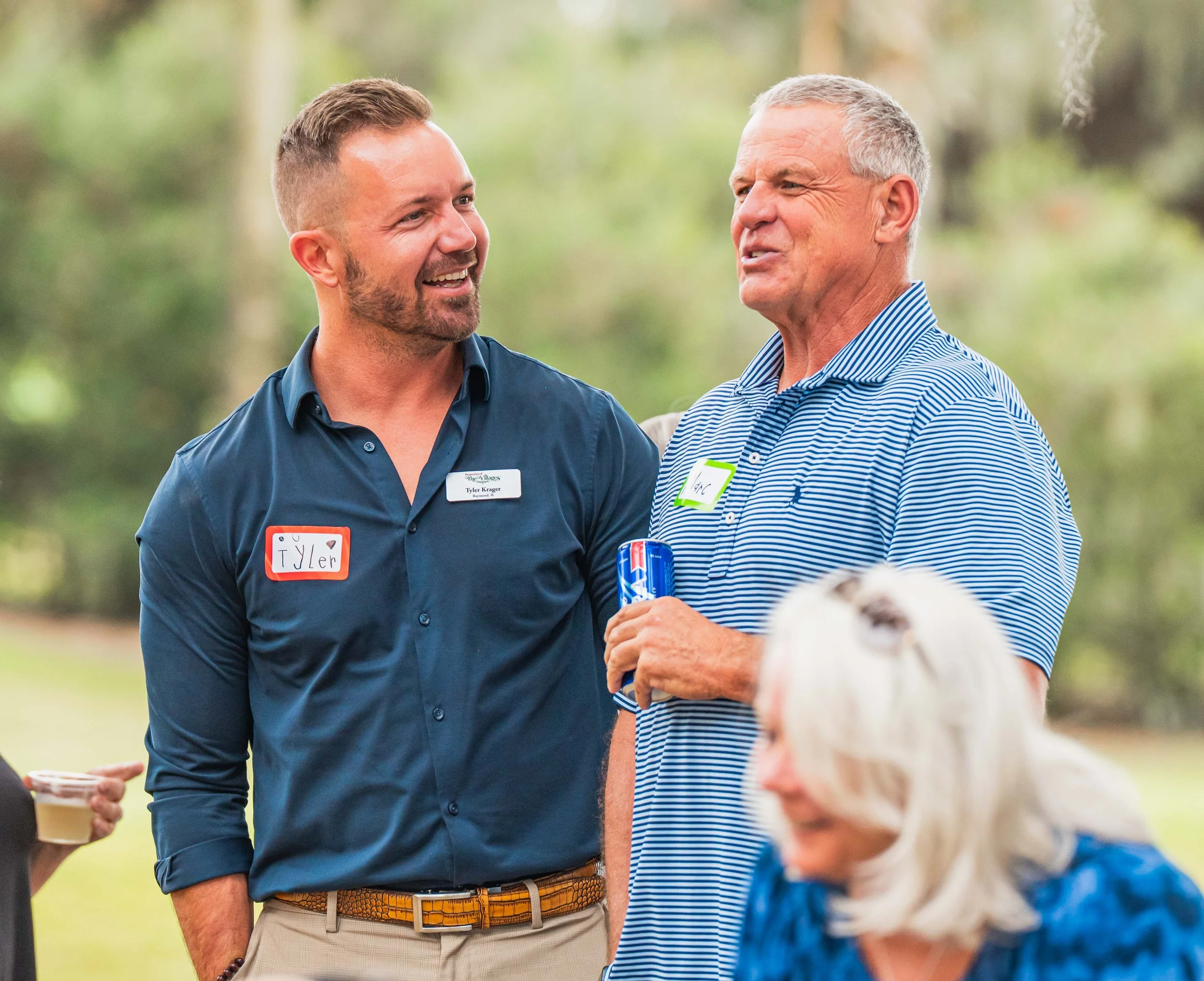 Two men engaged in conversation outdoors at a social gathering. One wears a navy blue shirt labeled 'TYLER'; the other wears a striped polo shirt and holds a can of soda.
