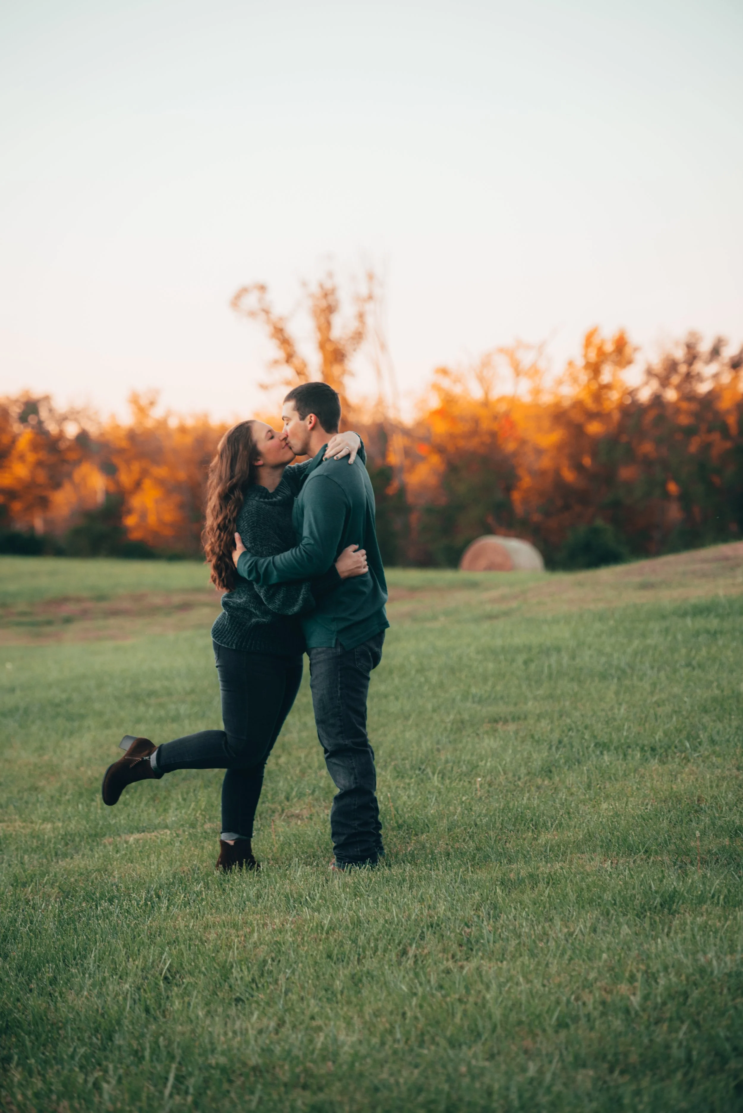 A couple kissing on a grassy field during sunset, with trees in autumn colors in the background.
