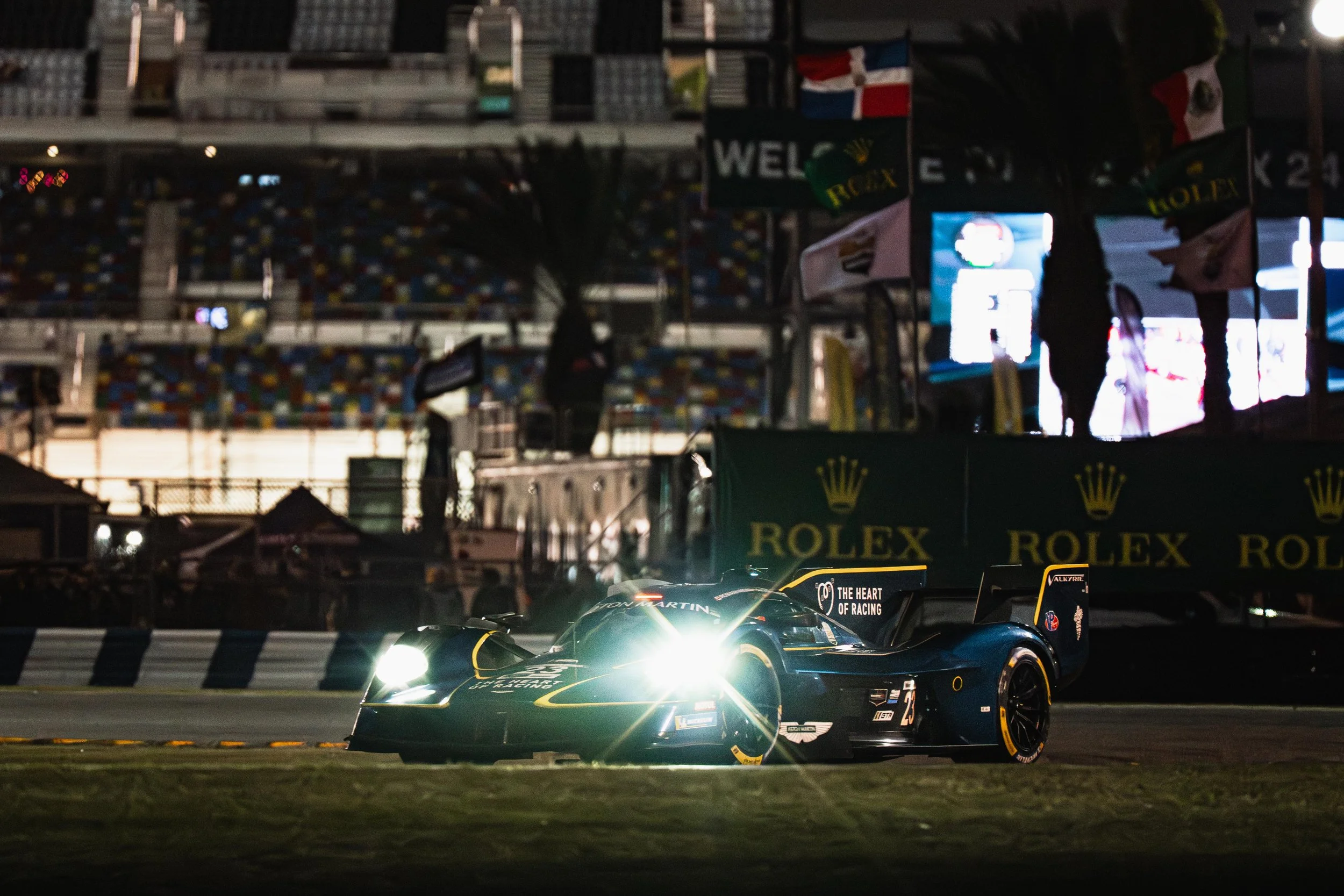 A race car with bright headlights on a race track at night, with flags and sponsor banners, including Rolex, visible in the background.