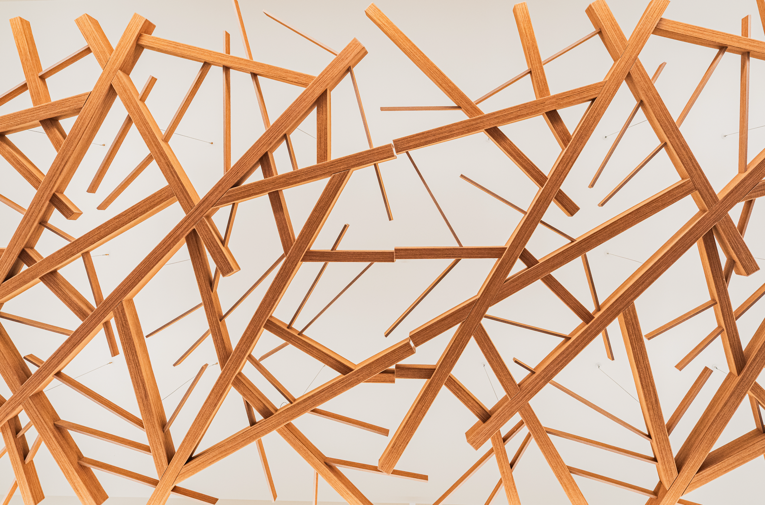 A modern ceiling installation made of numerous interconnected wooden beams forming an abstract, geometric pattern.