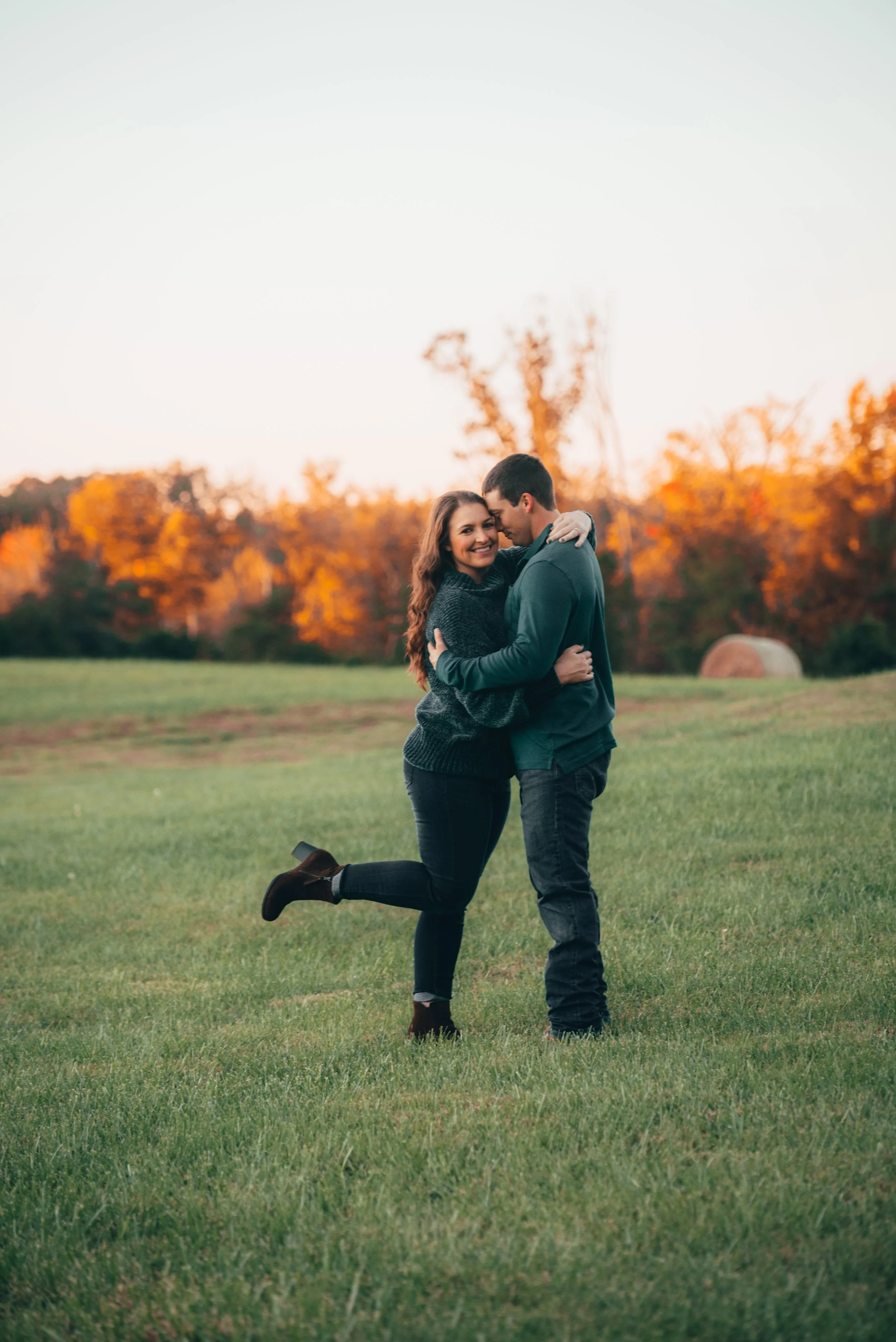 A happy couple embracing in a grassy field during fall, with autumn-colored trees and a hay bale in the background. Engagement photo session
