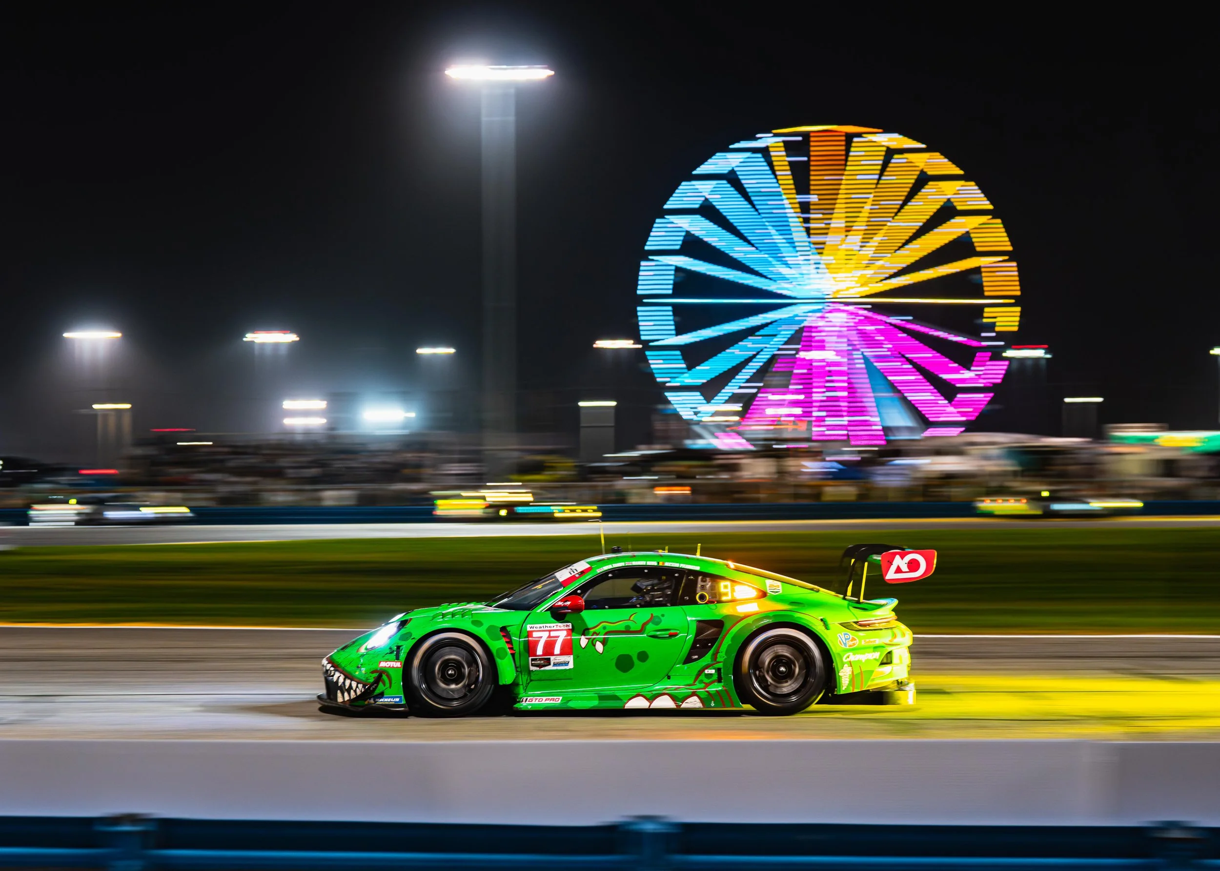 A green race car with a cartoonish face design on the front, speeding on a track at night with colorful Ferris wheel lights in the background.