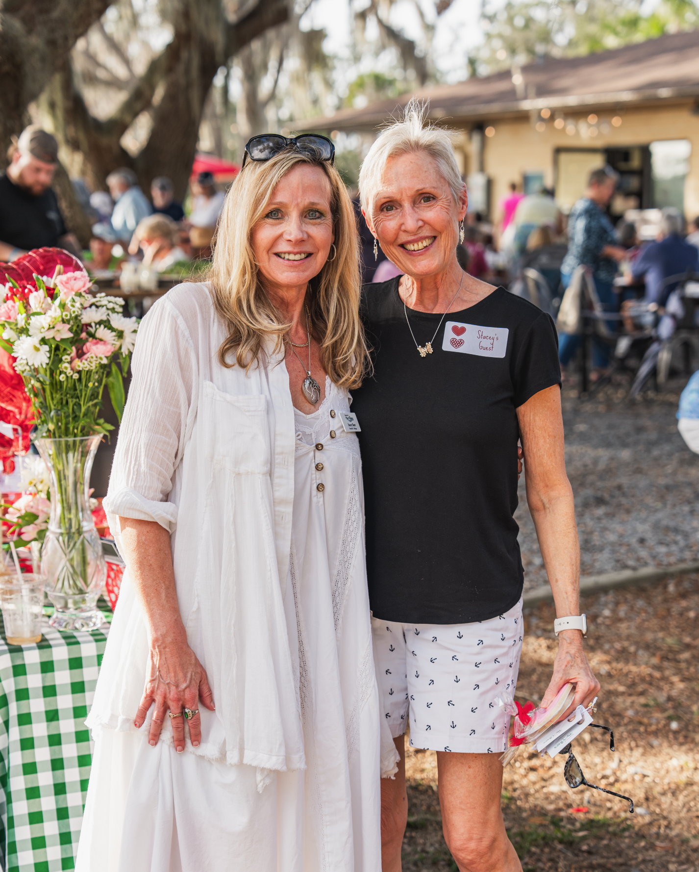 Two women standing outdoors at an event, smiling at the camera. One woman has long blonde hair, sunglasses on her head, and is wearing a white dress with a necklace. The other woman has short gray hair, is wearing a black t-shirt with a name tag and 