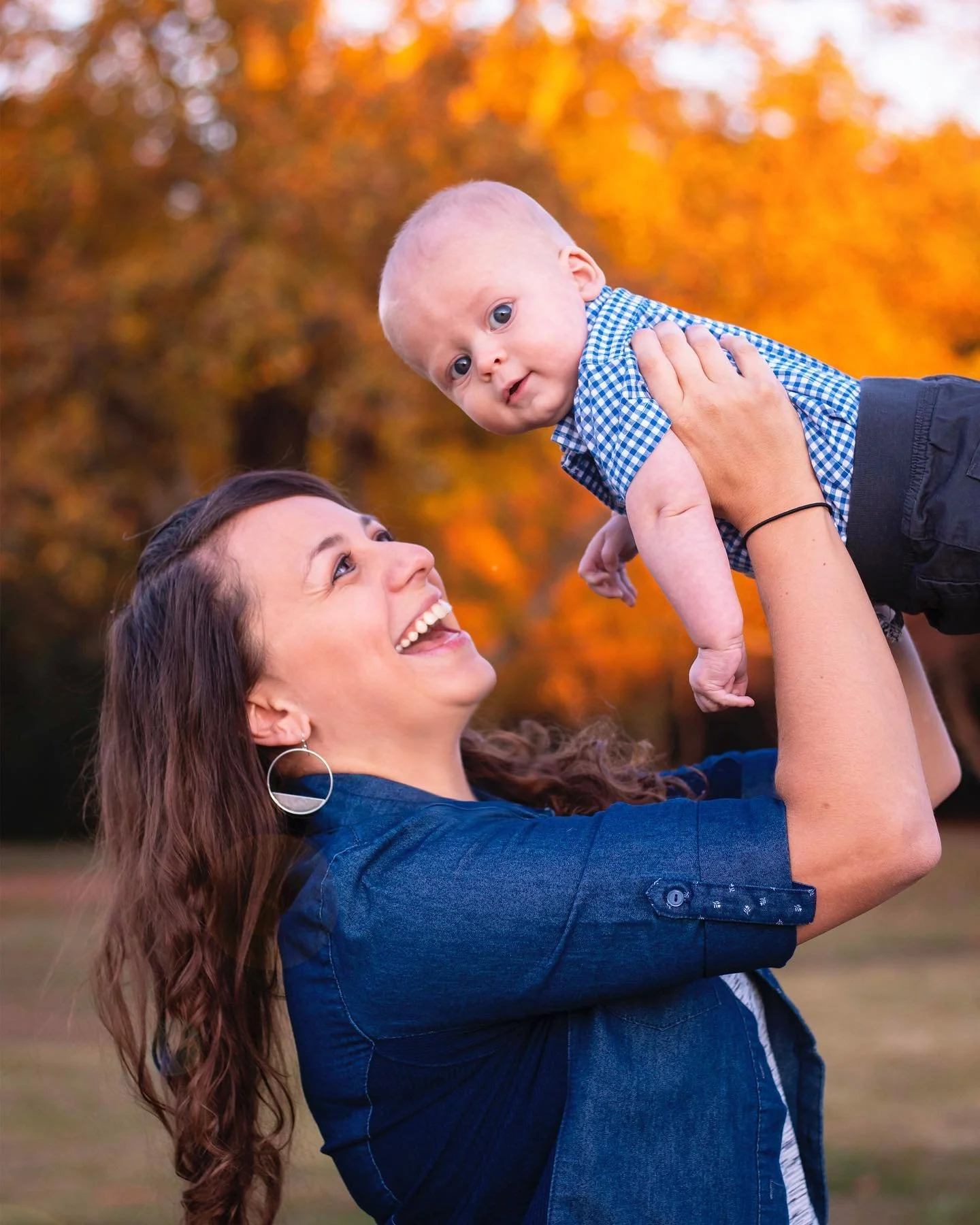 A woman with long curly hair and earrings smiling while holding a baby boy in the air outdoors during fall, with orange and yellow leaves in the background.