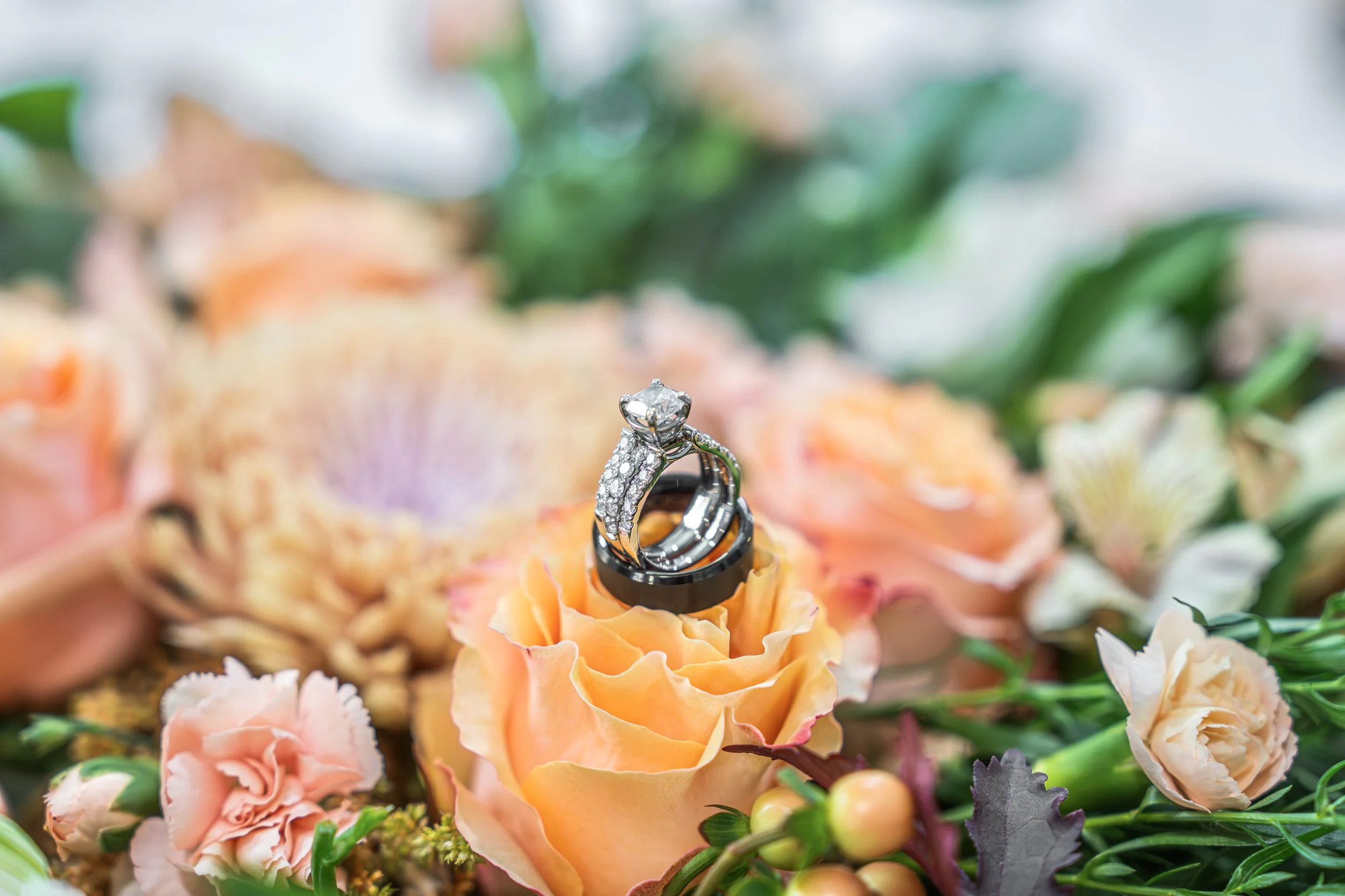 Engagement rings, a diamond solitaire and a band, are placed on a peach-colored rose surrounded by various flowers and greenery.