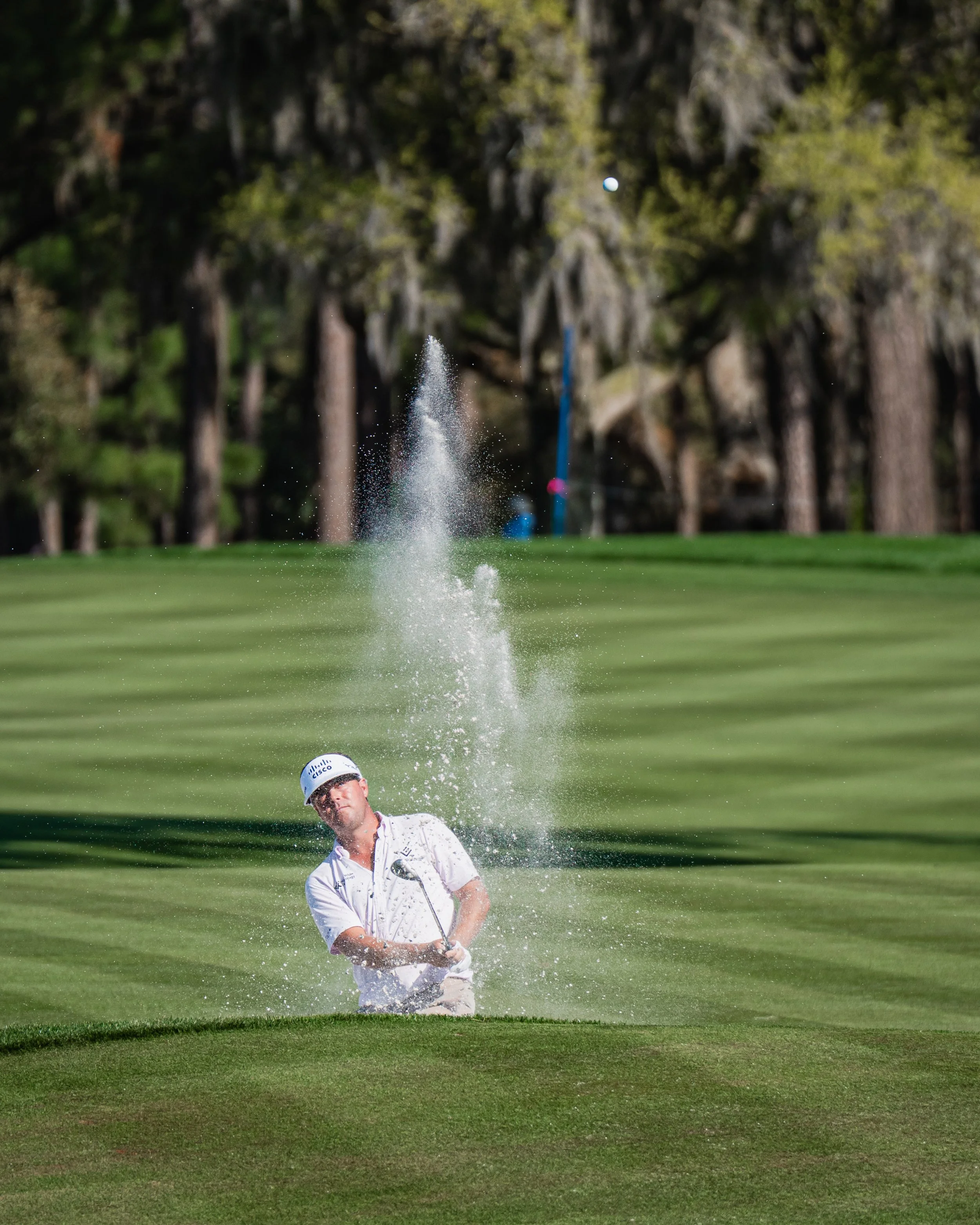 A man in golf attire, wearing a white cap and shirt, taking a shot from a sand trap on a golf course, with a golf ball in the air and a splash of sand.