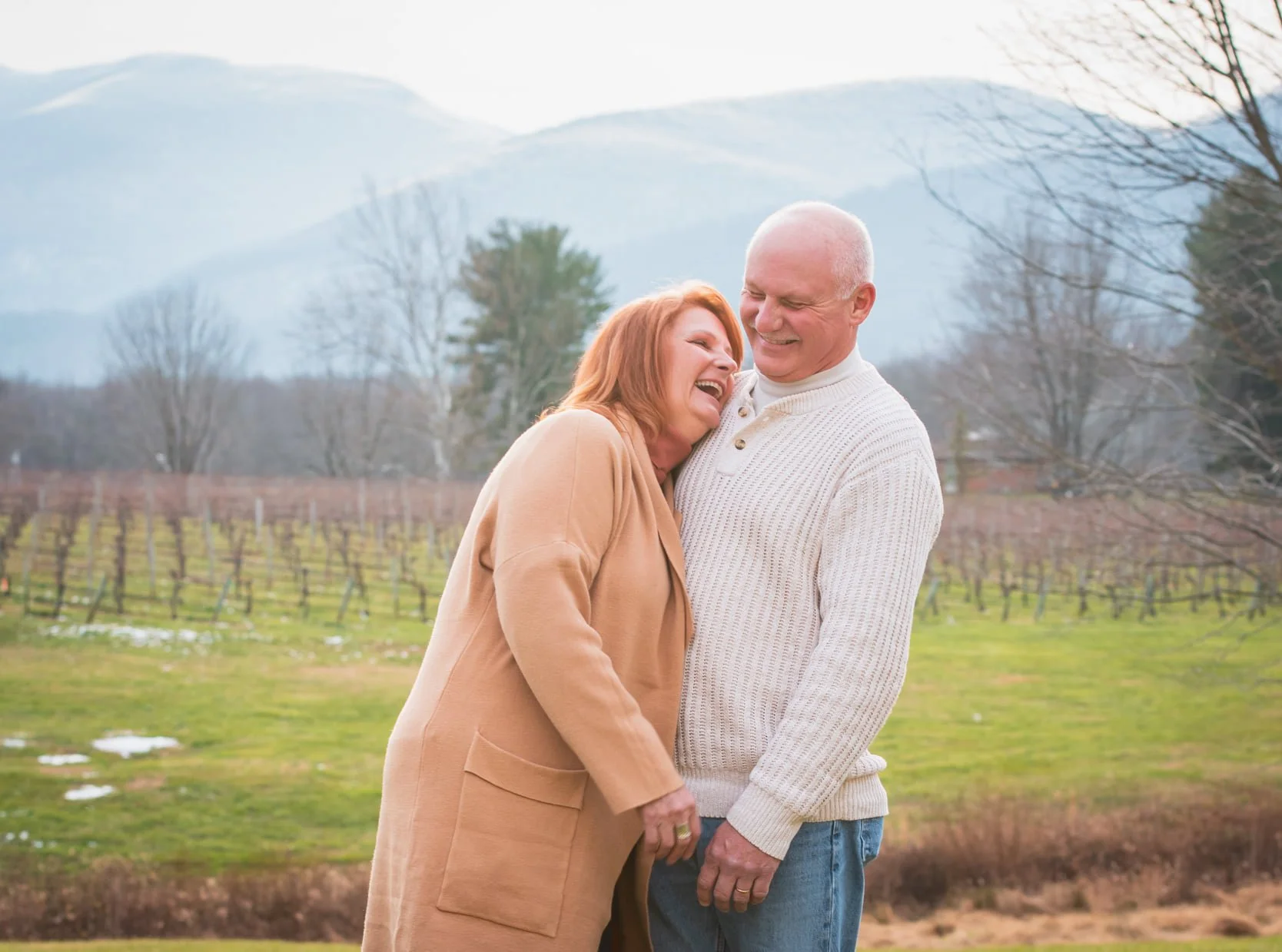 An elderly couple laughing and hugging outdoors in a vineyard with mountains in the background.