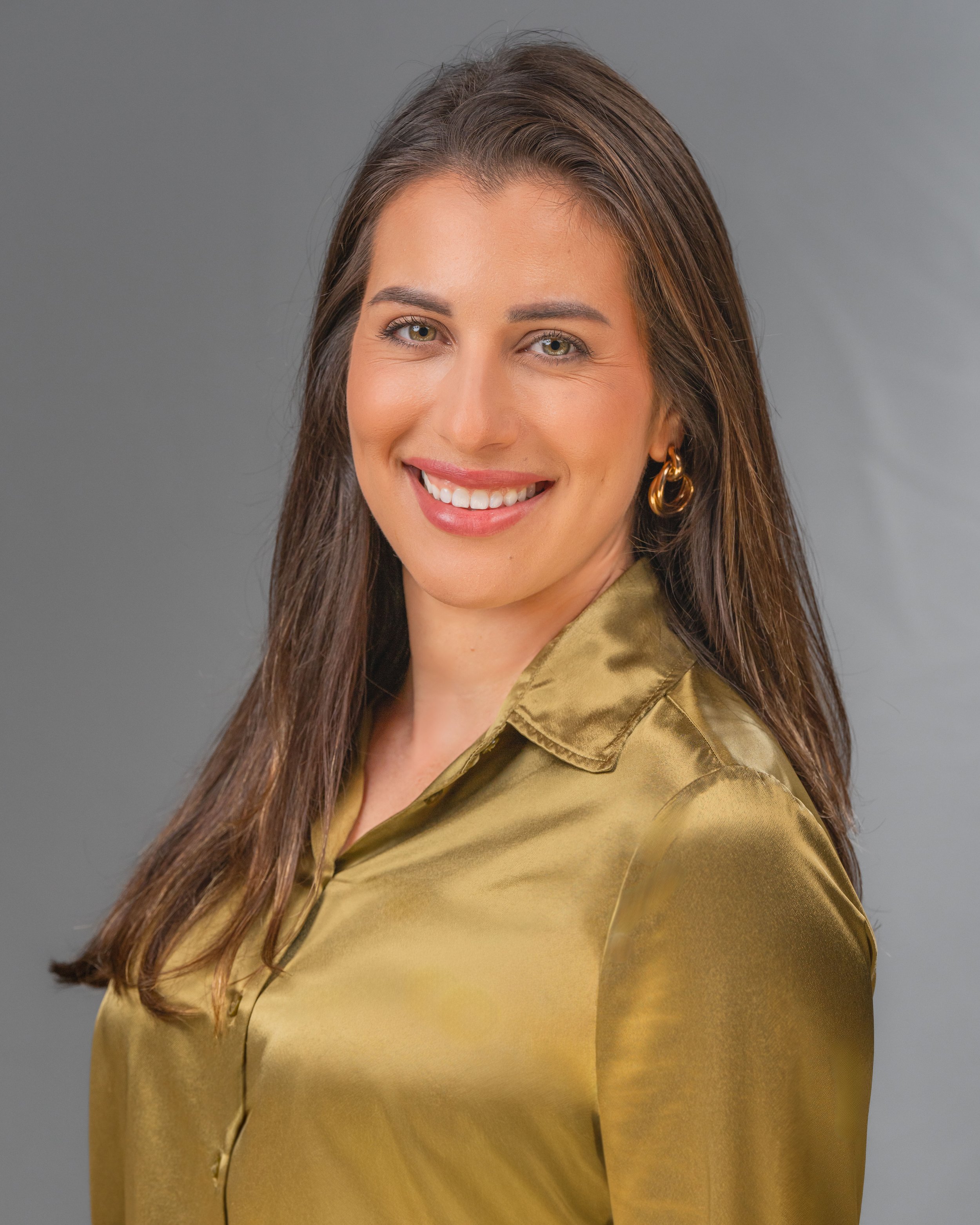Portrait photography, Central Florida. Corporate headshots. Headshot of a smiling woman with long brown hair, wearing gold earrings and an olive green satin shirt, against a gray background.