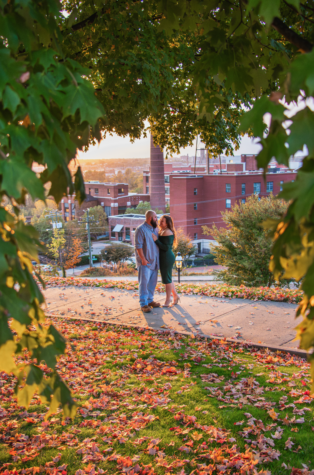 A couple stands on a sidewalk under a large tree with autumn leaves, overlooking a cityscape at sunset in Libby Hill, Richmond, Virginia