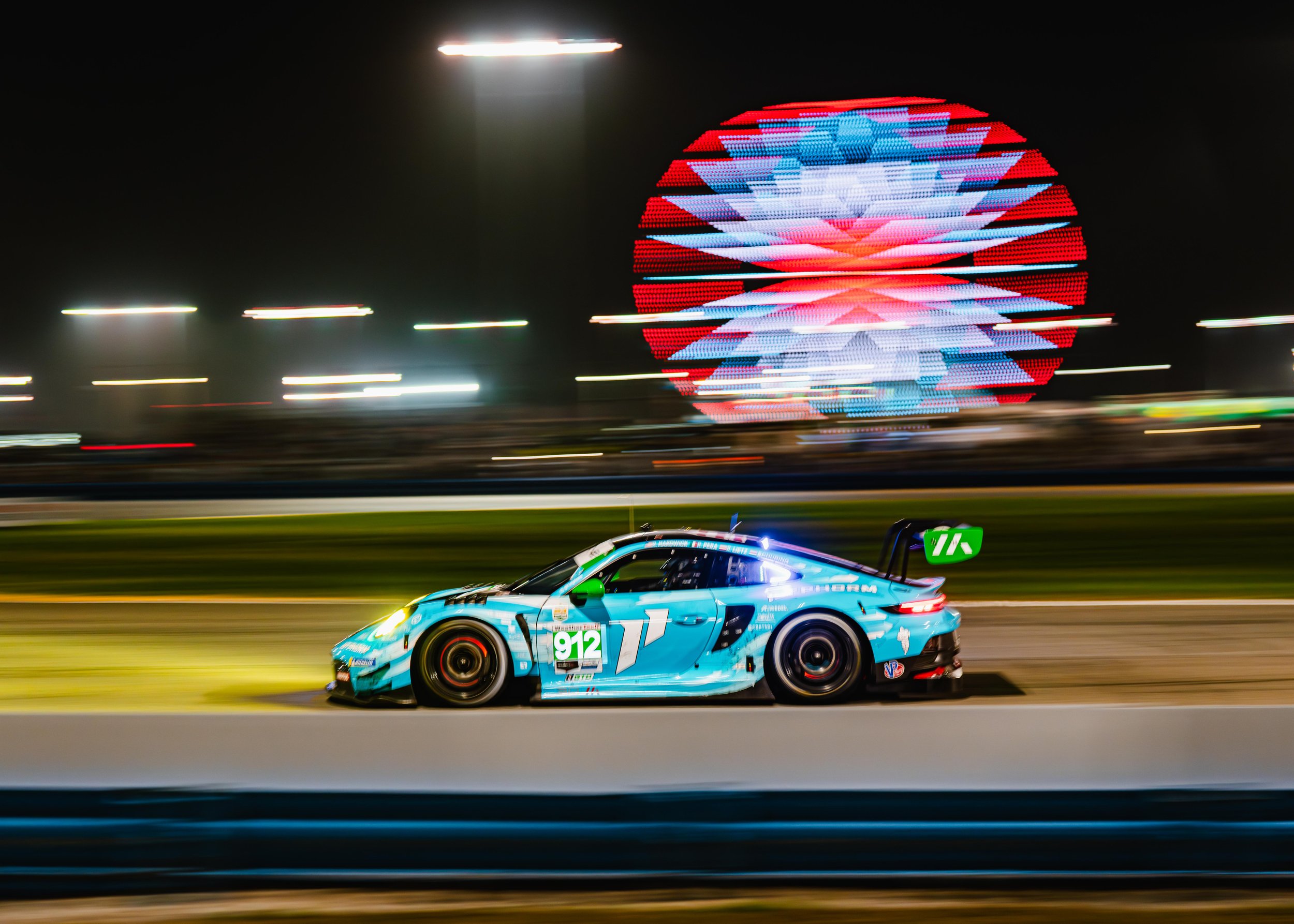 A blue race car, number 912, speeding on a race track at night with a blurred Ferris wheel illuminated in the background.