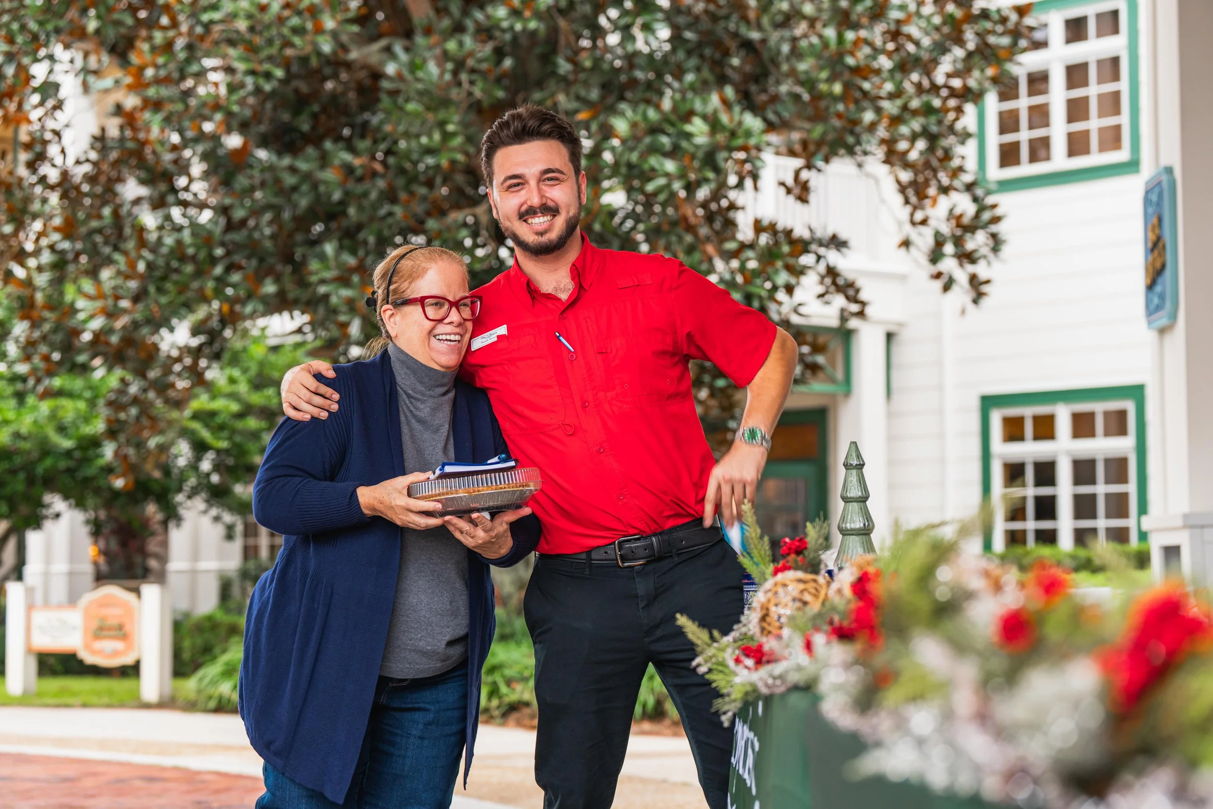 A smiling elderly woman with glasses holding a container of food, standing next to a young man in a red shirt with a nametag, outside in front of a white building with green trim, surrounded by trees and holiday decorations.