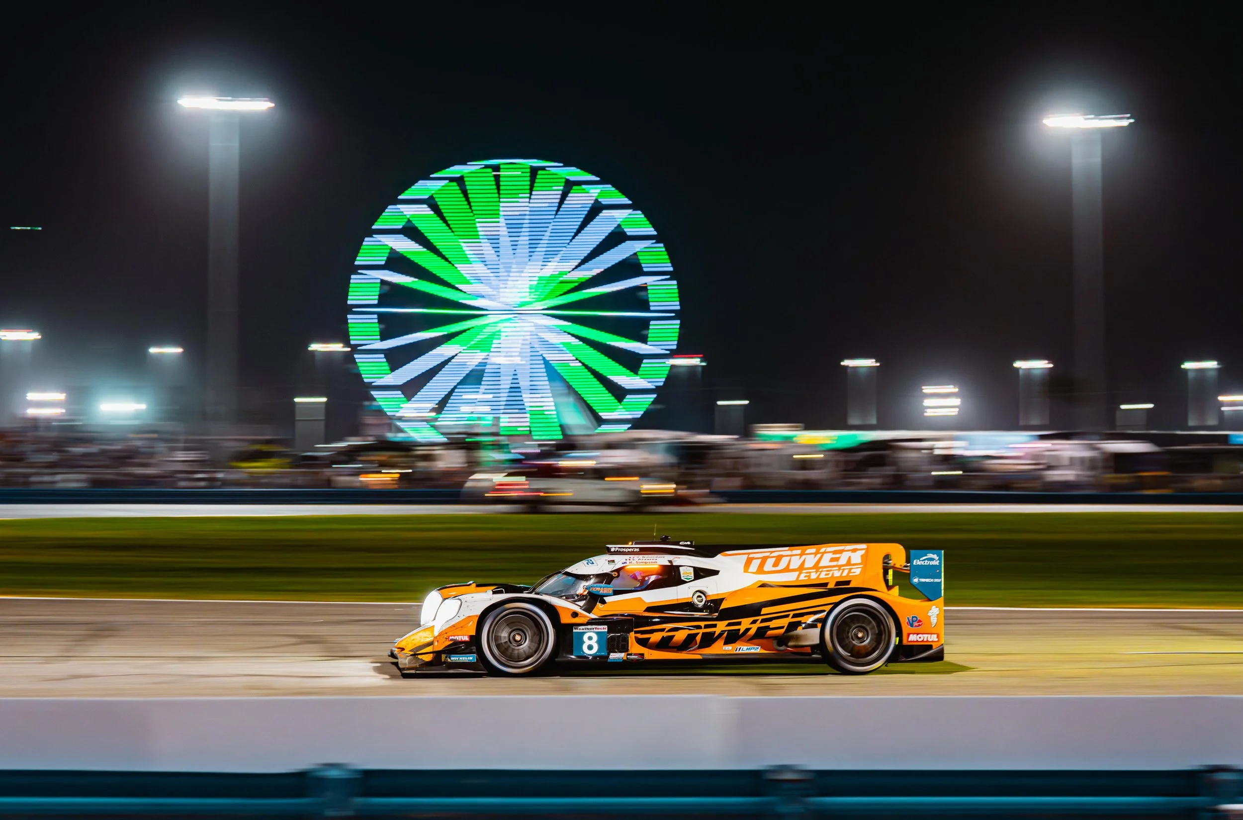 A race car driving on a track at night with a brightly lit Ferris wheel in the background.