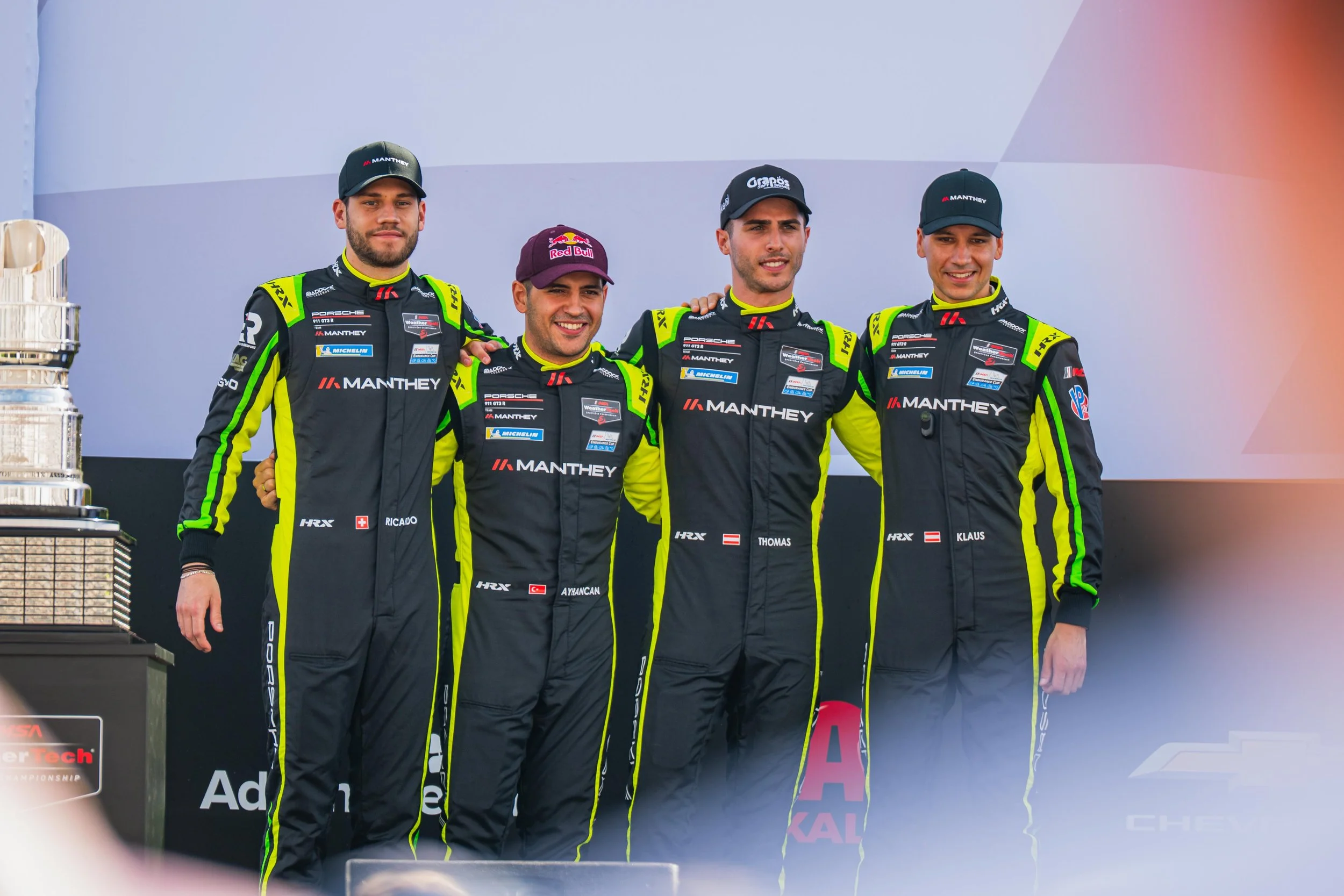 Four race car drivers standing on a podium with arms around each other, wearing black racing suits with green accents and hats, smiling after a motorsport race