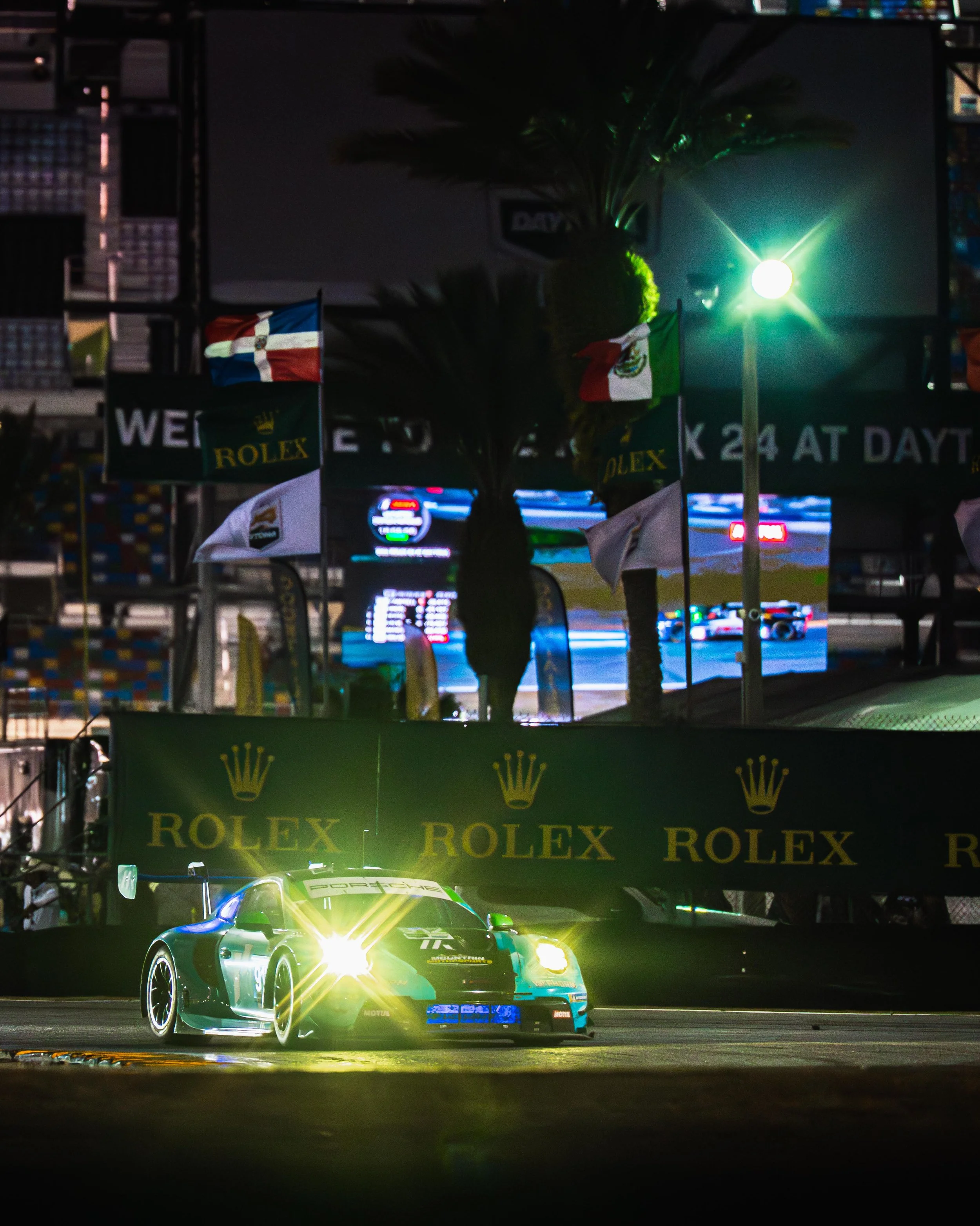 A race car on a track at night, illuminated by bright headlights. The background shows flags, palm trees, and digital screens with racing scenes, and banners advertising Rolex.