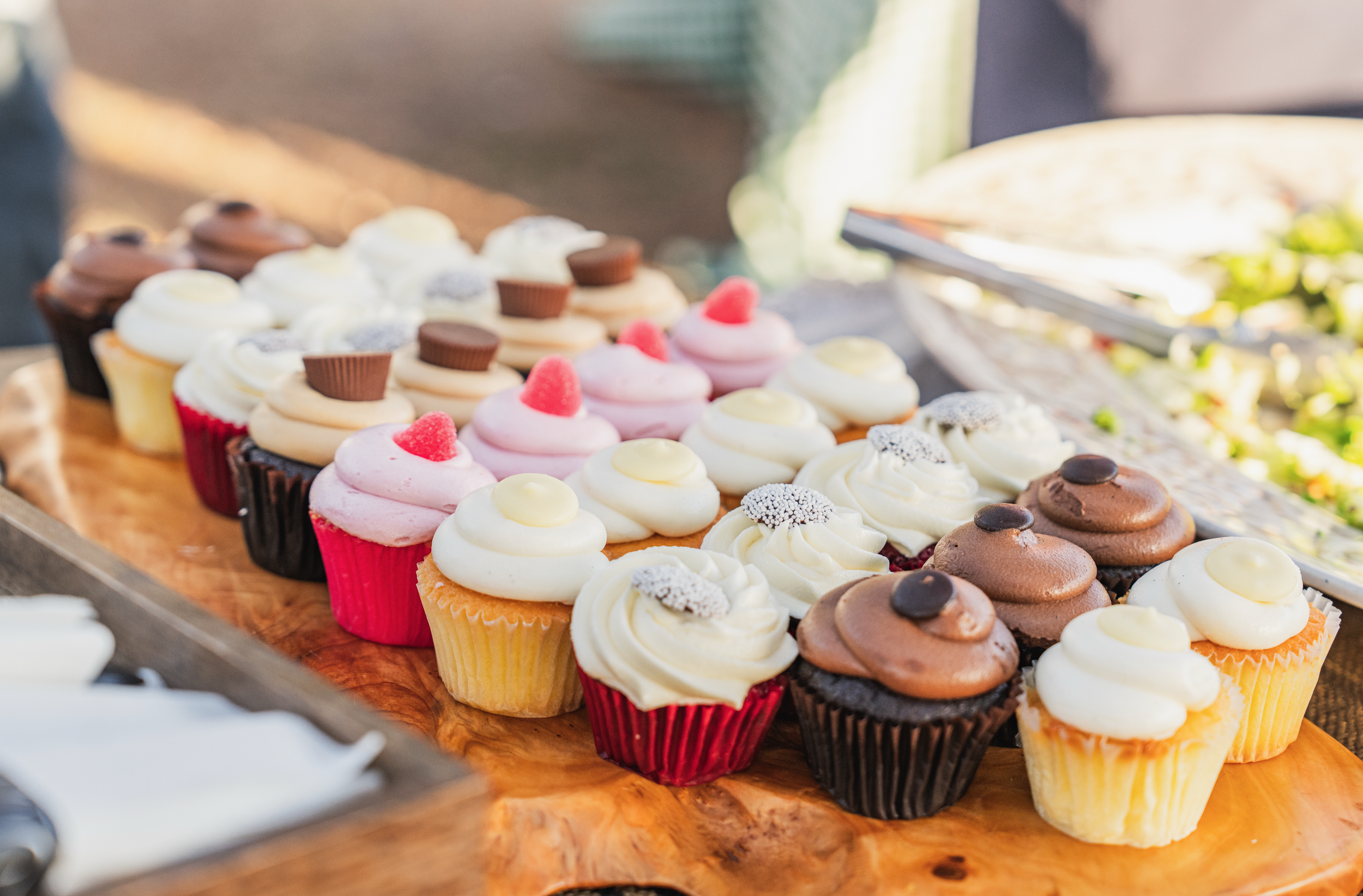 Assorted cupcakes with various toppings and frosting, displayed on a wooden platter at a bakery or cafe.
