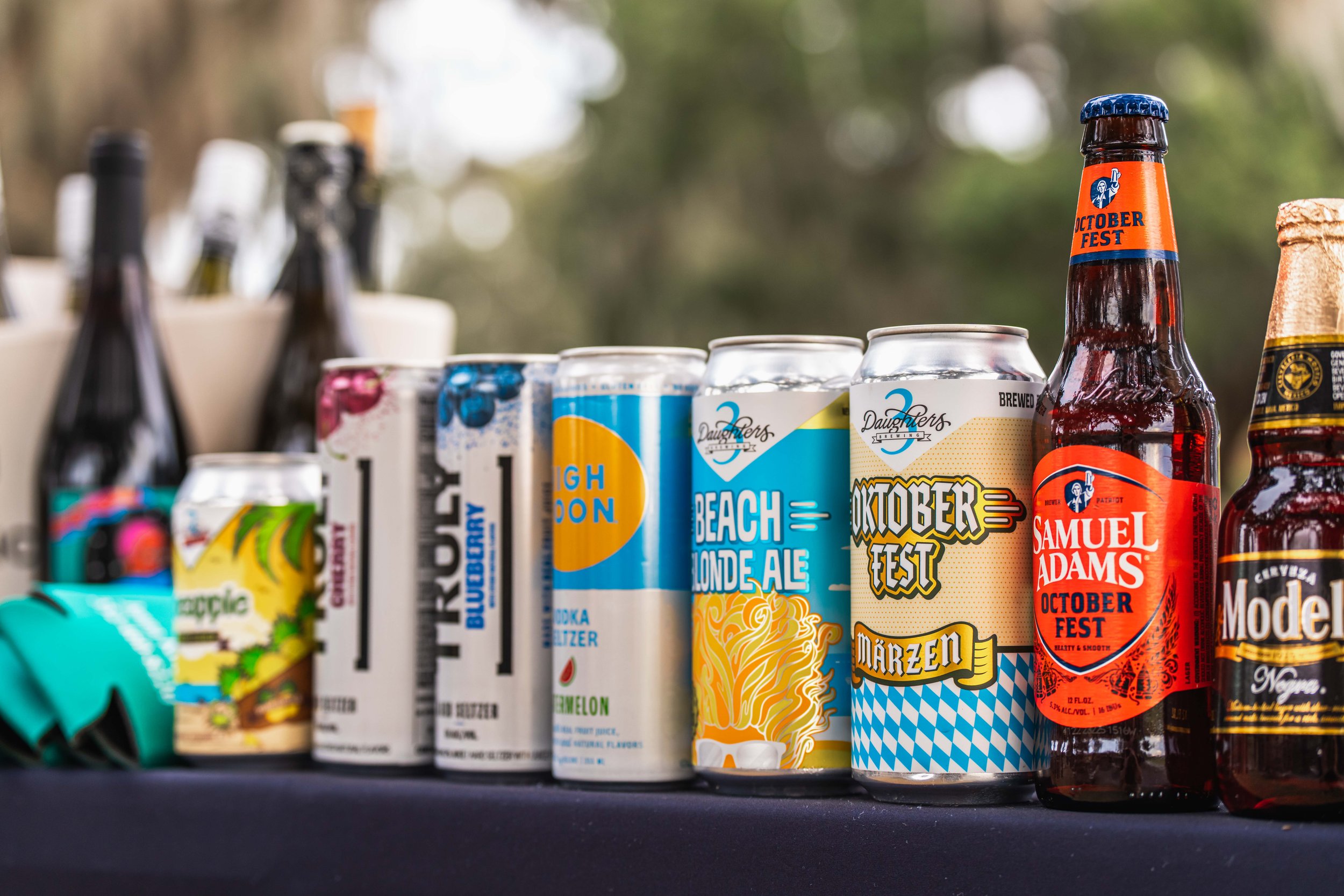 A variety of canned and bottled beverages on a black surface, including beers, seltzers, and cocktails, some with Oktoberfest and other labels, with bottles in the background.