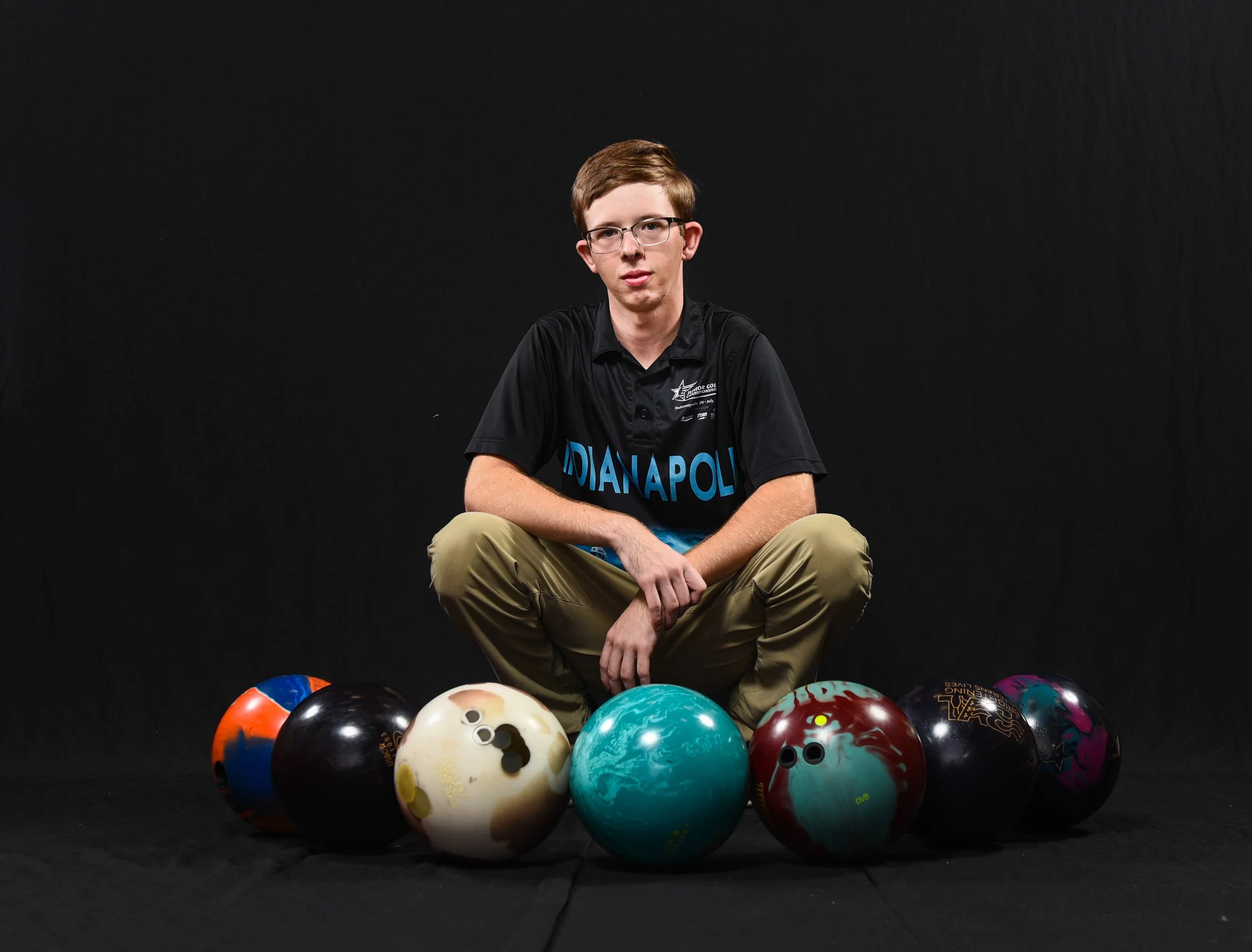 A young man with glasses and short hair, wearing a black Indianapolis bowling shirt, is sitting cross-legged on a black backdrop surrounded by seven colorful bowling balls. Senior photography
