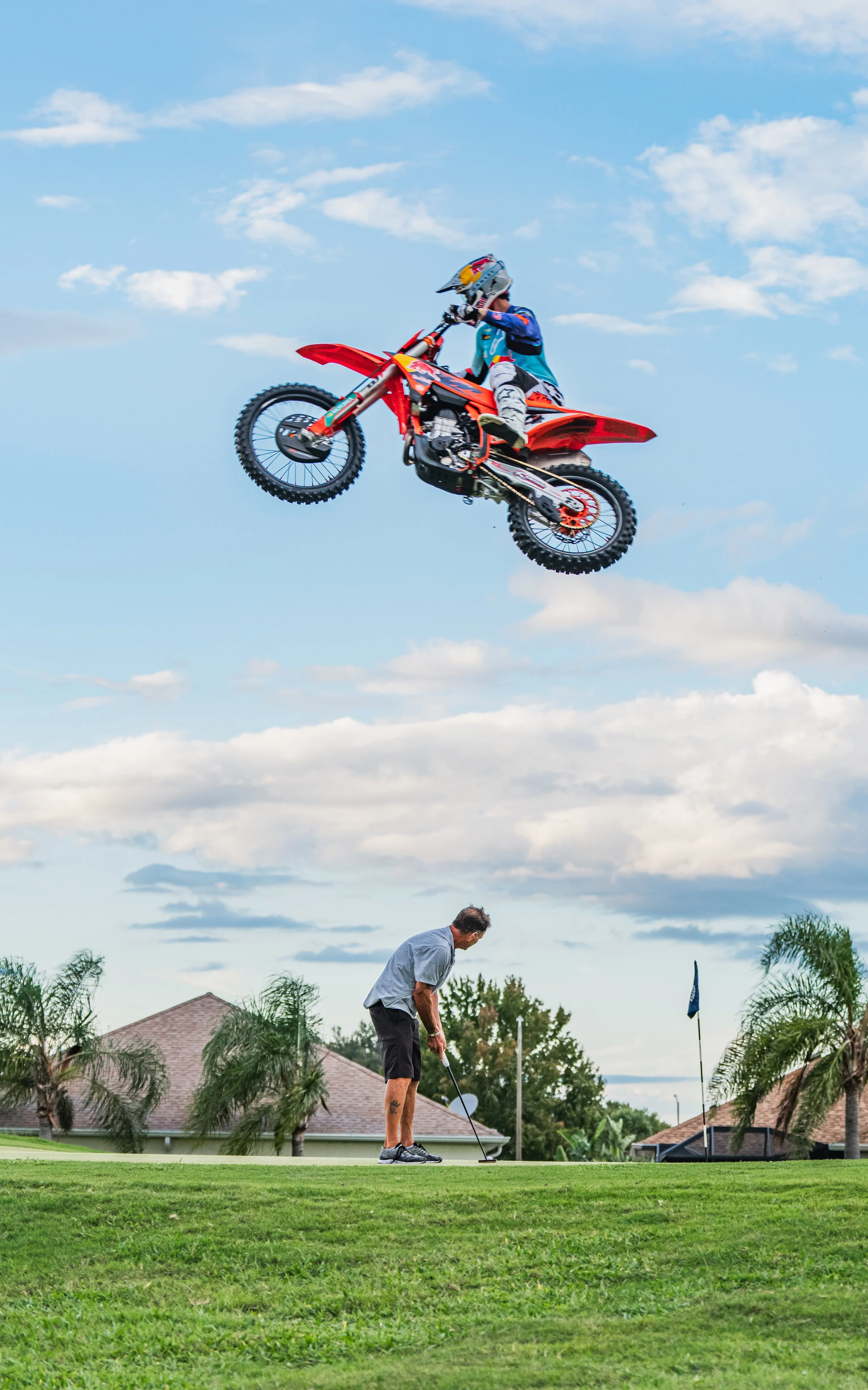 A person riding a red dirt bike is airborne in the sky, while another person prepares to putt on a golf course below.