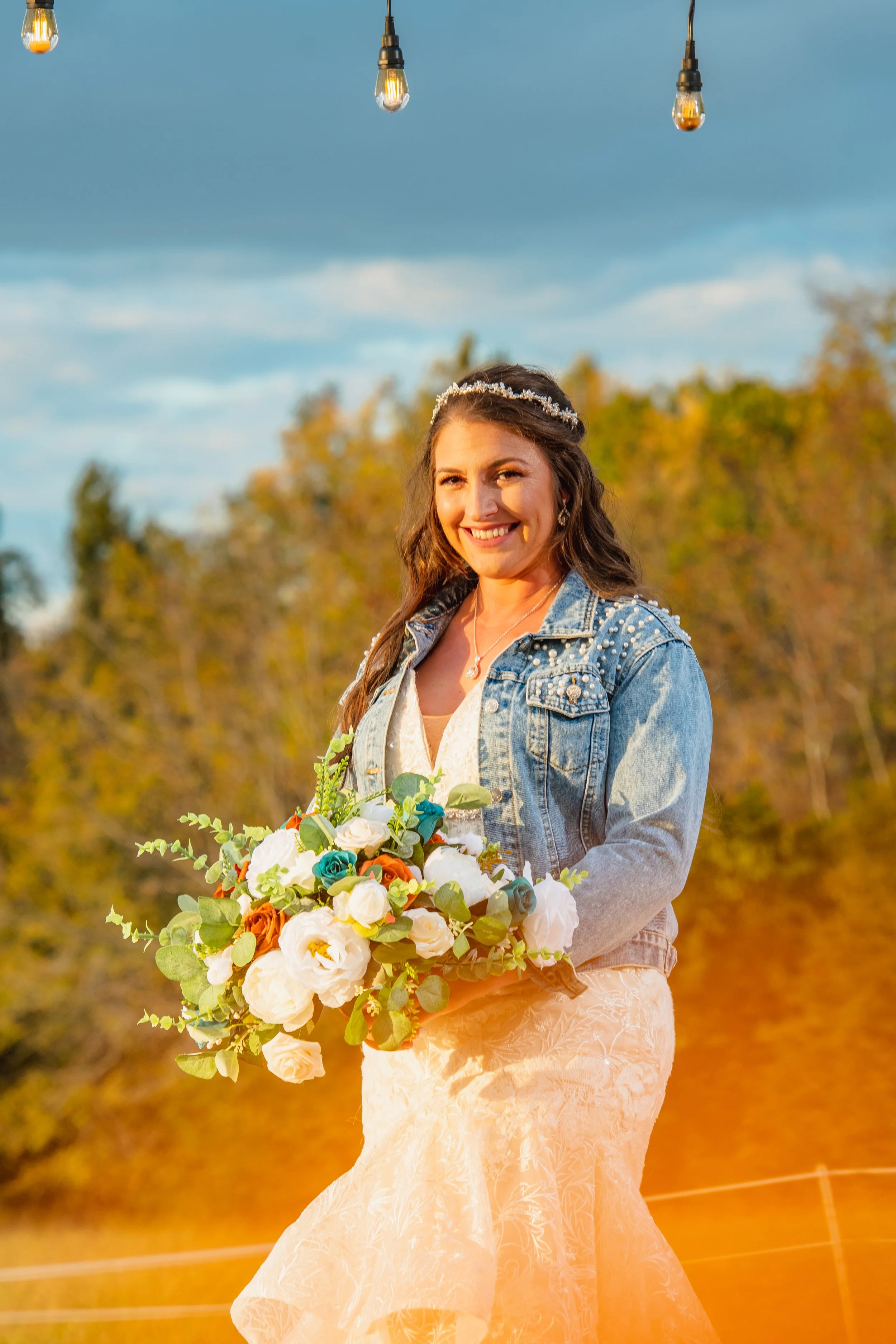 A bride in a wedding dress holding a bouquet of flowers, smiling outdoors during sunset, with string lights hanging above and trees in the background.