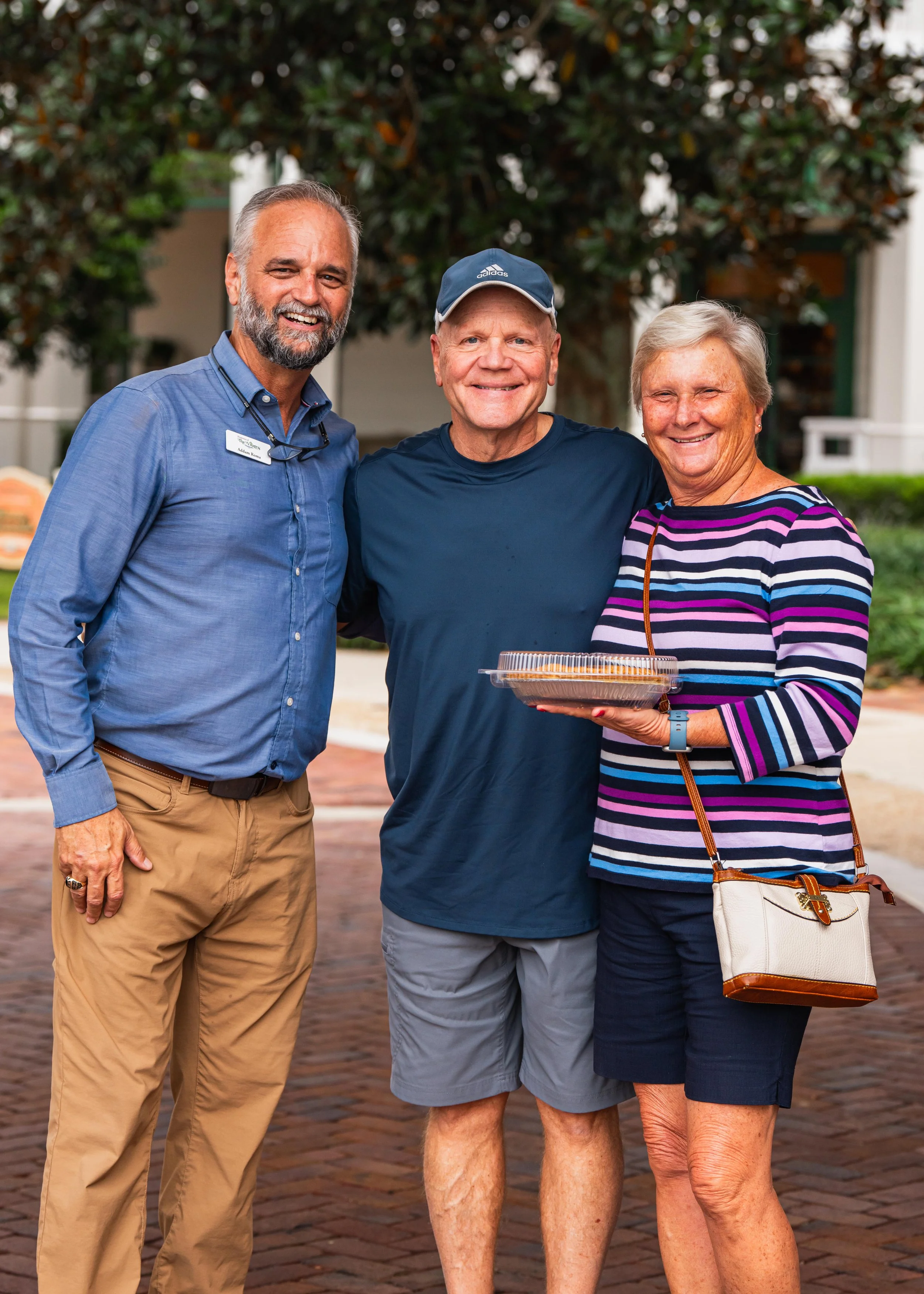 Three smiling adults standing outside on a brick path, with trees and buildings in the background. The woman on the right is holding a container of food.