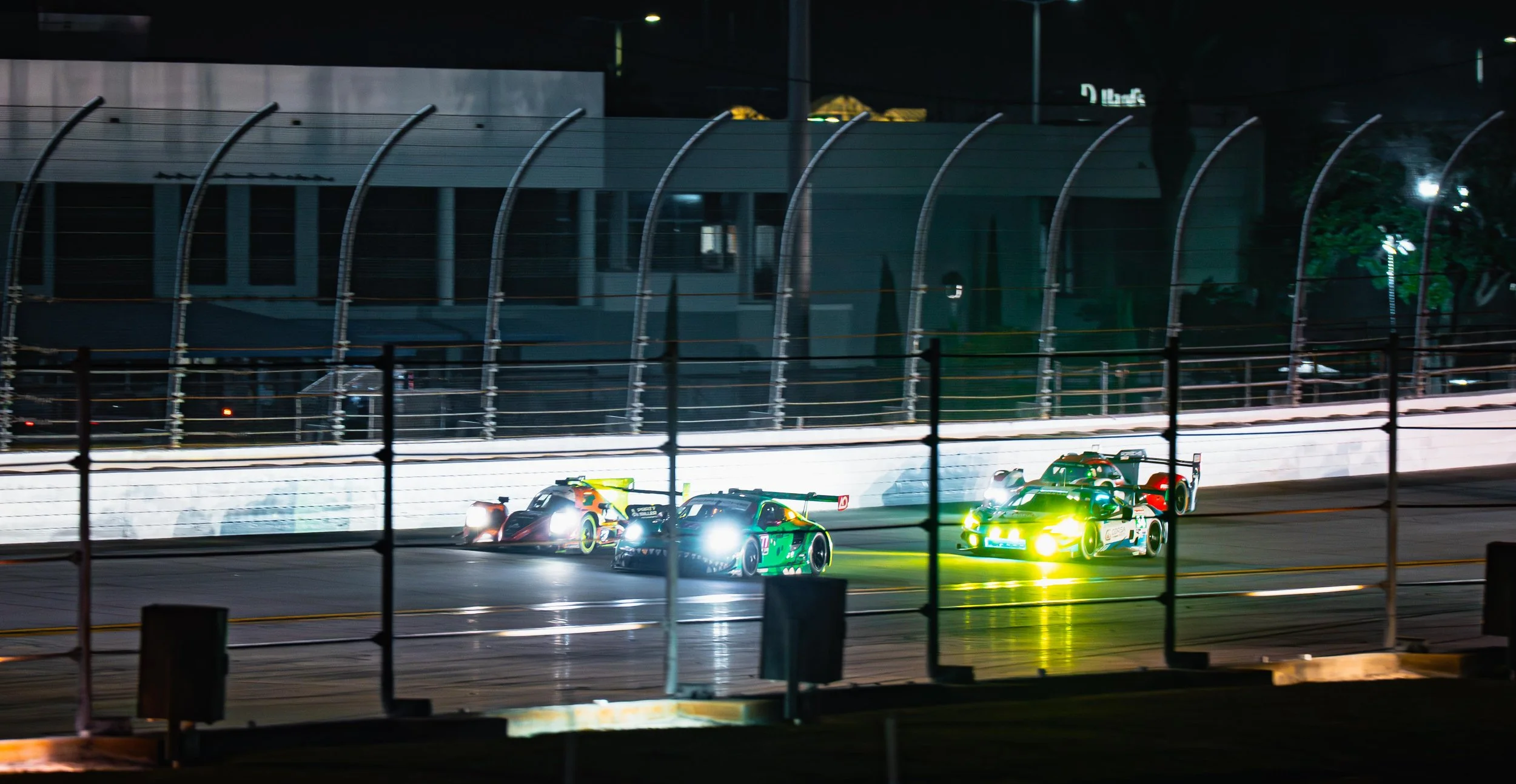 Nighttime race car competition on a track with four race cars illuminated by bright headlights, enclosed by a protective fence.