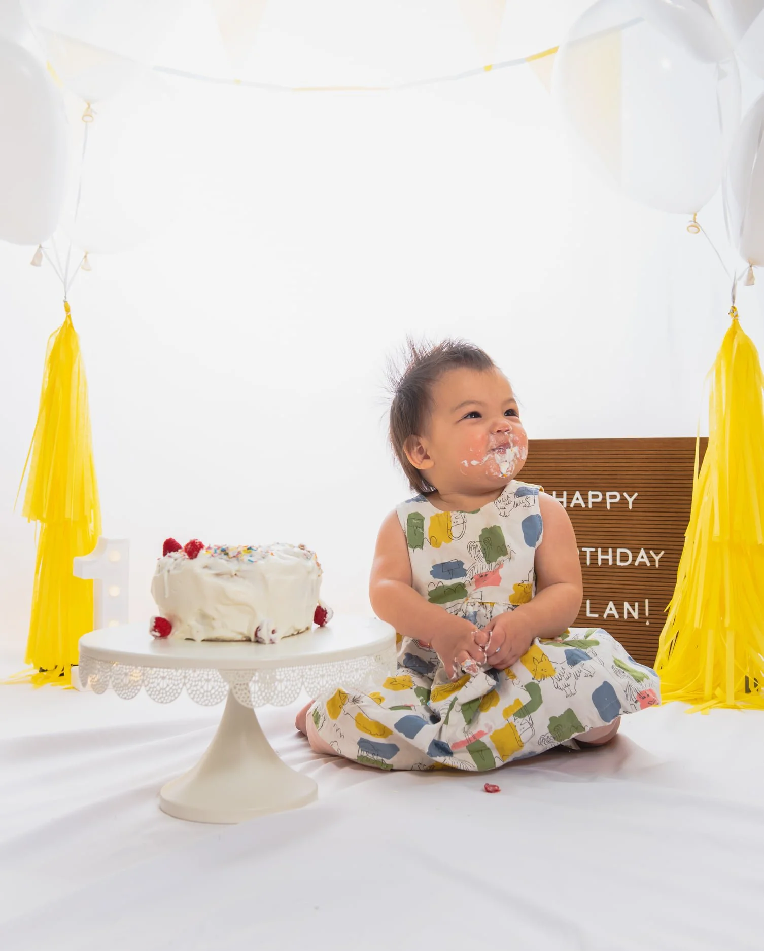 Cake smash photographer central Florida. A young child celebrating their first birthday with a cake, wearing a colorful dress, sitting on a white surface with balloons and yellow decorations around.